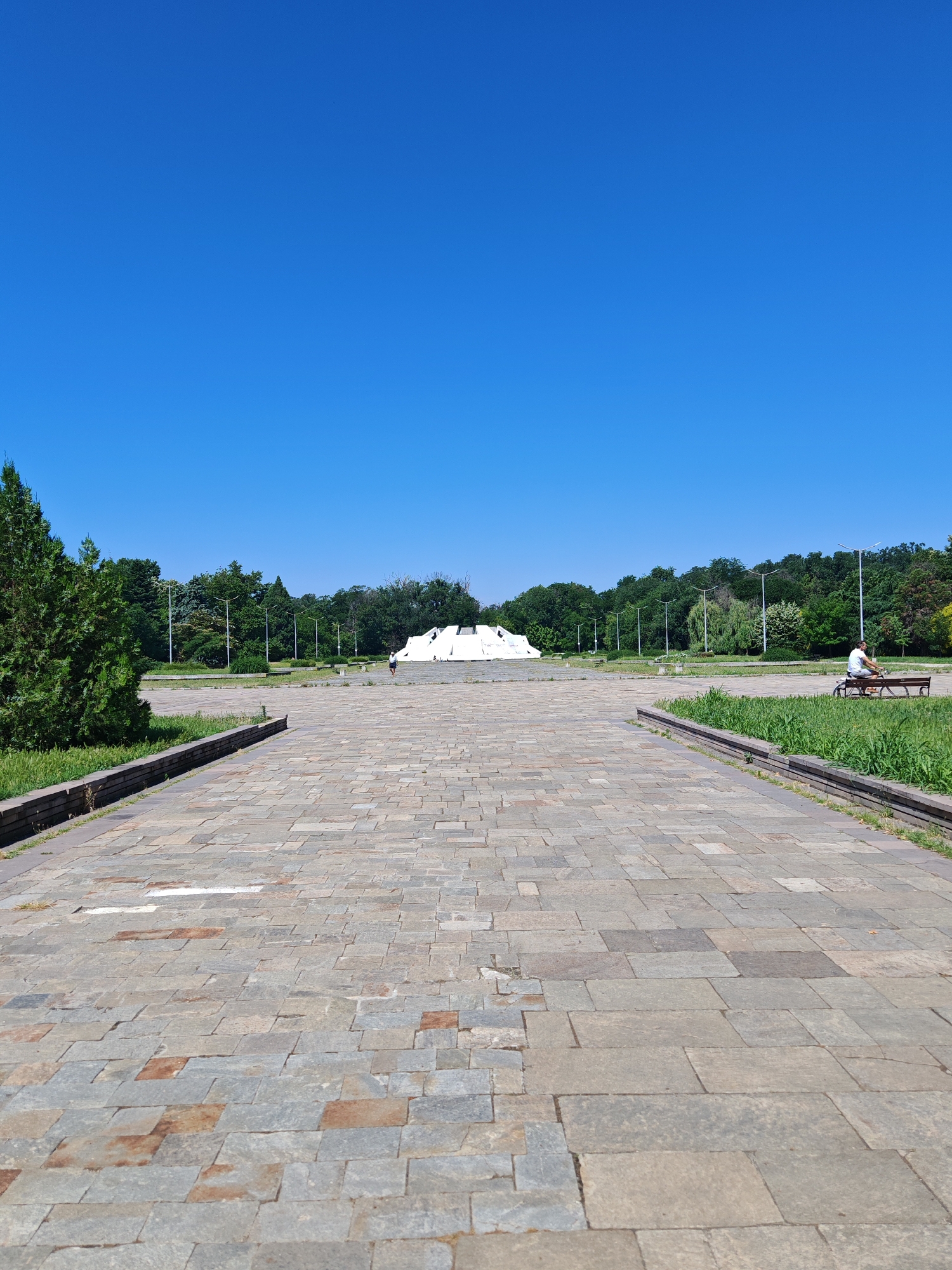 The image depicts a wide paved walkway leading towards a large, white, sculptural monument in the distance, situated in a park-like setting. The sky is a clear, vibrant blue. To the left of the walkway, there is a manicured green space with a large evergreen tree. To the right, another green space with grass and a bench is visible, occupied by a person sitting with a small dog. In the background, a dense line of trees forms a natural backdrop. The monument itself appears to be a large, abstract, white structure with angular lines. The walkway is paved with large, irregularly shaped stone tiles in shades of grey and brown. Streetlights are visible intermittently along the path in the background. This scene is located in the vicinity of the Alyosha Monument (Monument to the Soviet Soldier) in Plovdiv, Bulgaria, which is known for its expansive grounds and significant monument.