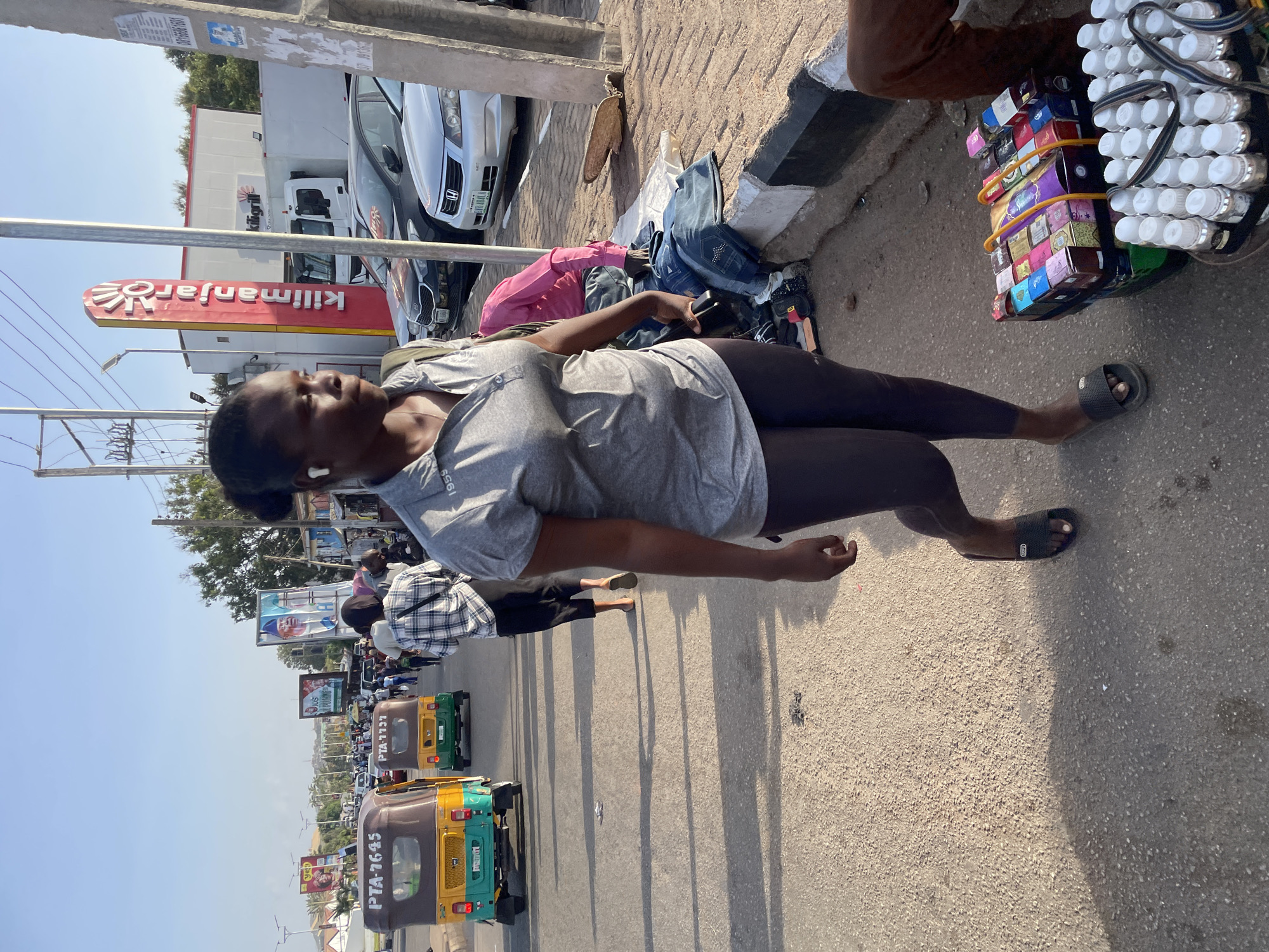 A street scene in Jos, Nigeria features a woman in a gray shirt and black leggings standing on a sidewalk next to what appears to be a vendor selling goods. The vendor's merchandise includes small boxes, possibly sweets or medicine, and containers with white caps. Behind the woman, a street is filled with "keke" vehicles (auto rickshaws), a common mode of transportation in the region. In the background, signs and billboards are visible along the street. A gas station with the Kilimanjaro sign is visible. Several cars are parked nearby.