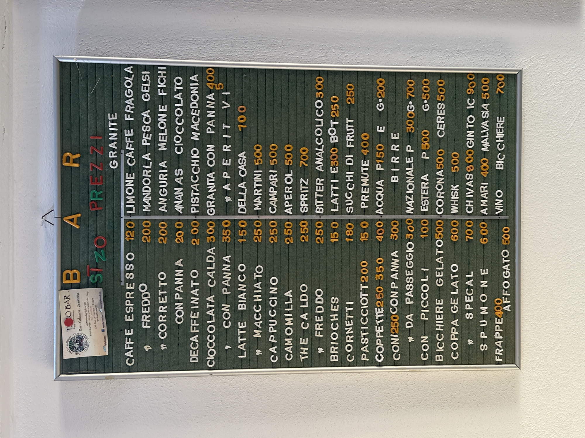 The image displays a dark green menu board with white, yellow, and red raised plastic letters, framed in metal and mounted on a textured white wall. The top left corner features a small business card or sticker for "TOOBAR Bar - Gelateria - Graniteria" located in "Canneto-Lipari (Me)-Italia".
The board lists "PREZZI" (Prices) for various items. Beverages include "CAFFE ESPRESSO" (120), "FREDDO" (200), "CORRETTO" (200), "CON PANNA" (200), "DECAFFEINATO" (200), "CIOCCOLATA CALDA" (300), "CON PANNA" (350), "LATTE BIANCO" (150), "MACCHIATO" (250), "CAPPUCCINO" (250), "CAMOMILLA" (250), "THE CALDO" (250), and "FREDDO" (250).
Granita flavors are listed as "LIMONE CAFFE FRAGOLA," "MANDORLA PESCA GELSI," "ANGURIA MELONE FICHI," "ANANAS CIOCCOLATO," and "PISTACCHIO MACEDONIA," with "GRANITA CON PANNA" priced at 400.
"APERITIVI" include "DELLA CASA" (700), "MARTINI" (500), "CAMPARI" (500), "APEROL" (500), "SPRITZ" (700), "BITTER ANALCOLICO" (300), "LATTI" (300), "BOT" (250), "SUCCHI DI FRUTT" (250), and "PREMUTE" (400). "ACQUA" is 150 (P) and 200 (G).
"BIRRE" options are "NAZIONALE" (P 300, G 700), "ESTERA" (P 500, G 500), "CORONA" (500), "CERES" (500). Other alcoholic drinks include "WHISK" (500), "CHIVAS" (800), "GIN TO" (900), "AMARI" (400), "MALVA SIA" (500), and "VINO BICCHIERE" (700).
Food items include "BRIOCHES" (150), "CORNETTI" (180), "PASTICCIOTT" (200), "COPPETTE" (250, 350, 400), "CON PANNA" (300), "DA PASSEGGIO" (300), "CON PICCOLI" (100), "BICCHIERE GELATO" (500), "COPPA GELATO" (600), "SPECAL" (700), "SPUMONE" (600), "FRAPPE" (400), and "AFFOGATO" (500).