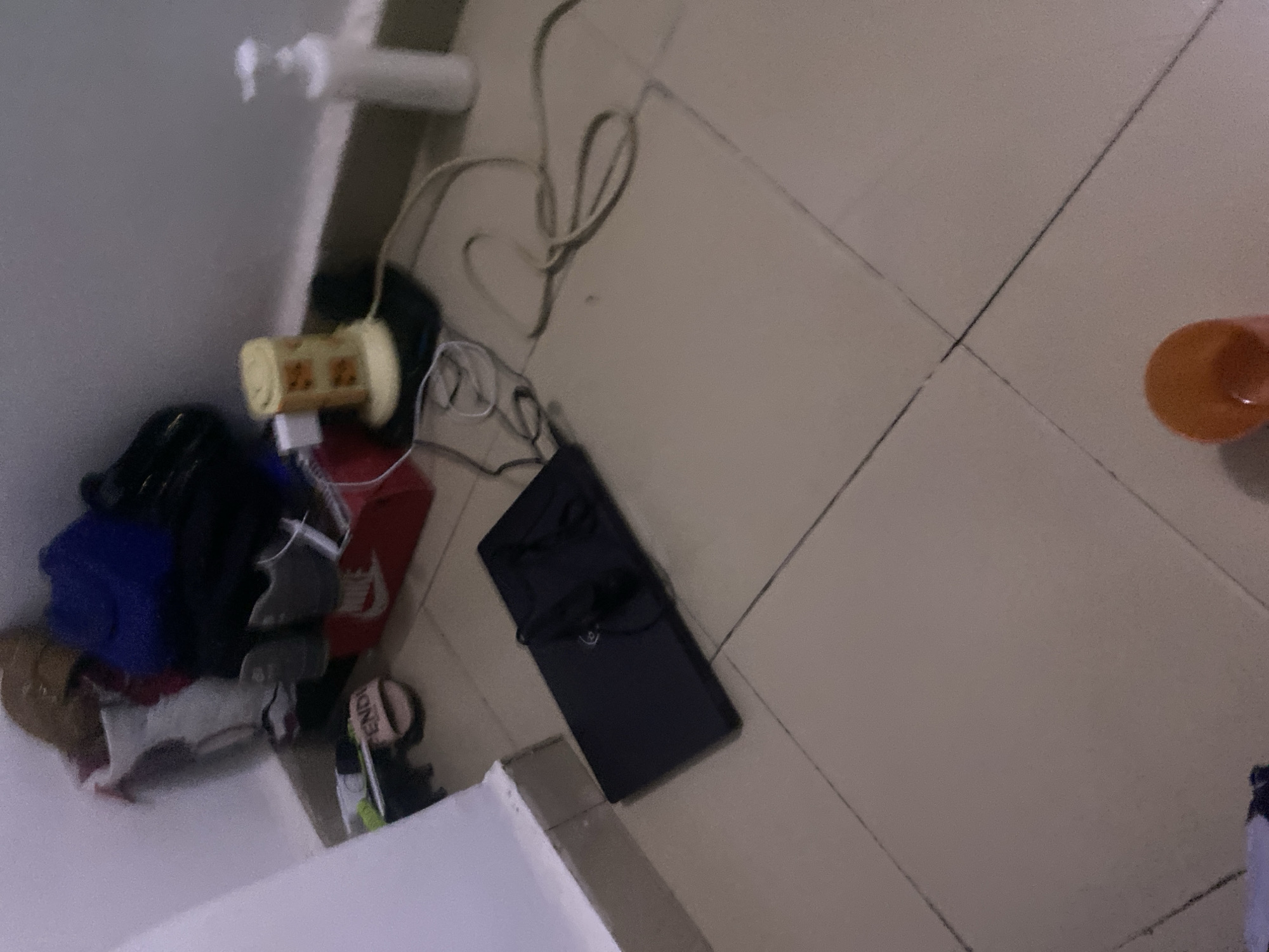 The image depicts a cluttered corner of a room, possibly a closet or a storage area, with items haphazardly placed on the floor and against the wall. In the background, there is a stack of items including a blue duffel bag, a white and red Nike shoe box, and what appears to be a pair of grey sneakers. To the left, a pair of pink slides with the word "FENDI" is visible. In the mid-ground, a black laptop is laid flat on the tiled floor, with a pair of black earbuds placed on top of it. To the right of the laptop, a white dispenser bottle, possibly hand soap or lotion, stands upright. Adjacent to it, a cylindrical object with a yellow and orange pattern, likely a humidifier or air freshener, is plugged into a white adapter. The floor is covered in square beige tiles with dark grout lines. The scene appears to be in a domestic setting. There is no indication of the city of Howayi, Nigeria, or any specific location within it.