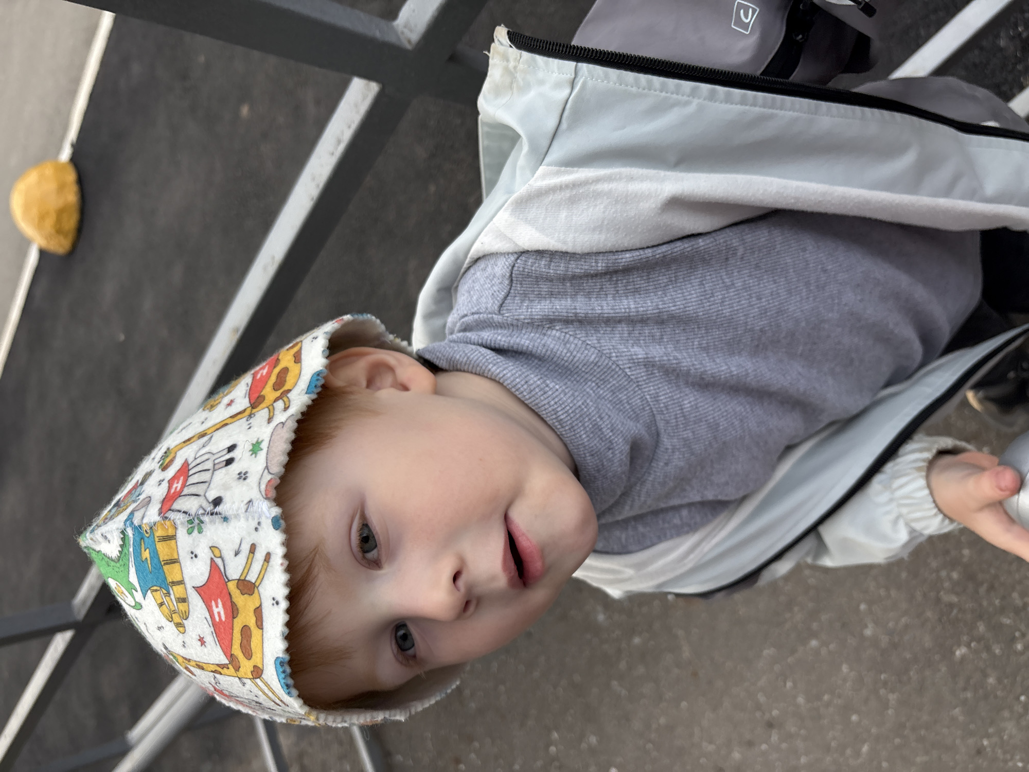 A young child, approximately 2-4 years old, with fair skin, light reddish-brown hair, and light-colored eyes, is depicted looking slightly upwards and to the viewer's left, with lips slightly parted, in an outdoor setting. The child wears a white, conical-shaped hat with a scalloped brim, patterned with various colorful cartoon animals including giraffes and zebras, some featuring a red 'H' symbol. Below the hat, the child is dressed in a grey ribbed turtleneck shirt and a light-colored, possibly light grey or off-white, hooded windbreaker jacket, worn open. The child's right hand is visible in the lower left quadrant of the image, grasping a metallic, cylindrical object. The immediate foreground and background consist of a dark grey paved surface, likely asphalt or a rubberized playground material, exhibiting light-colored specks. A grey metal railing or barrier extends horizontally in the mid-background, supported by a vertical post on the right side. Further in the background, a distinct bright yellow, dome-shaped object rests on the paved surface, accompanied by a narrow white line. The overall lighting indicates an outdoor daylight environment. The visual evidence does not provide identifying features to pinpoint a specific location within Izhevsk, Russia.