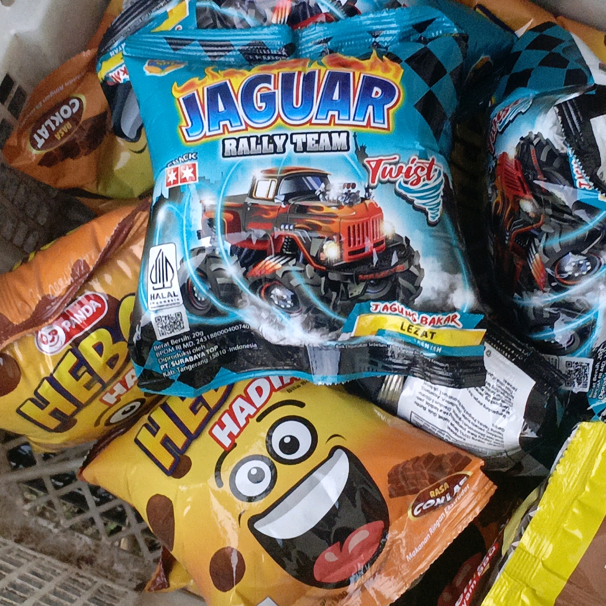 The image shows a close-up of several snack food packages piled in a basket, likely in Salatiga, Indonesia, as indicated by the text on some of the packaging. The prominent packaging is a bright blue bag featuring a monster truck with flames and the text "JAGUAR RALLY TEAM TWIST". Other visible packages include one with the "PANDA" logo and the text "HEB" in large yellow letters, and another orange package with the text "RASA COKLAT" (Chocolate Flavor). The lighting suggests an indoor or shaded outdoor environment. There are no people visible in the image, and the focus is entirely on the arrangement of the snack food items.