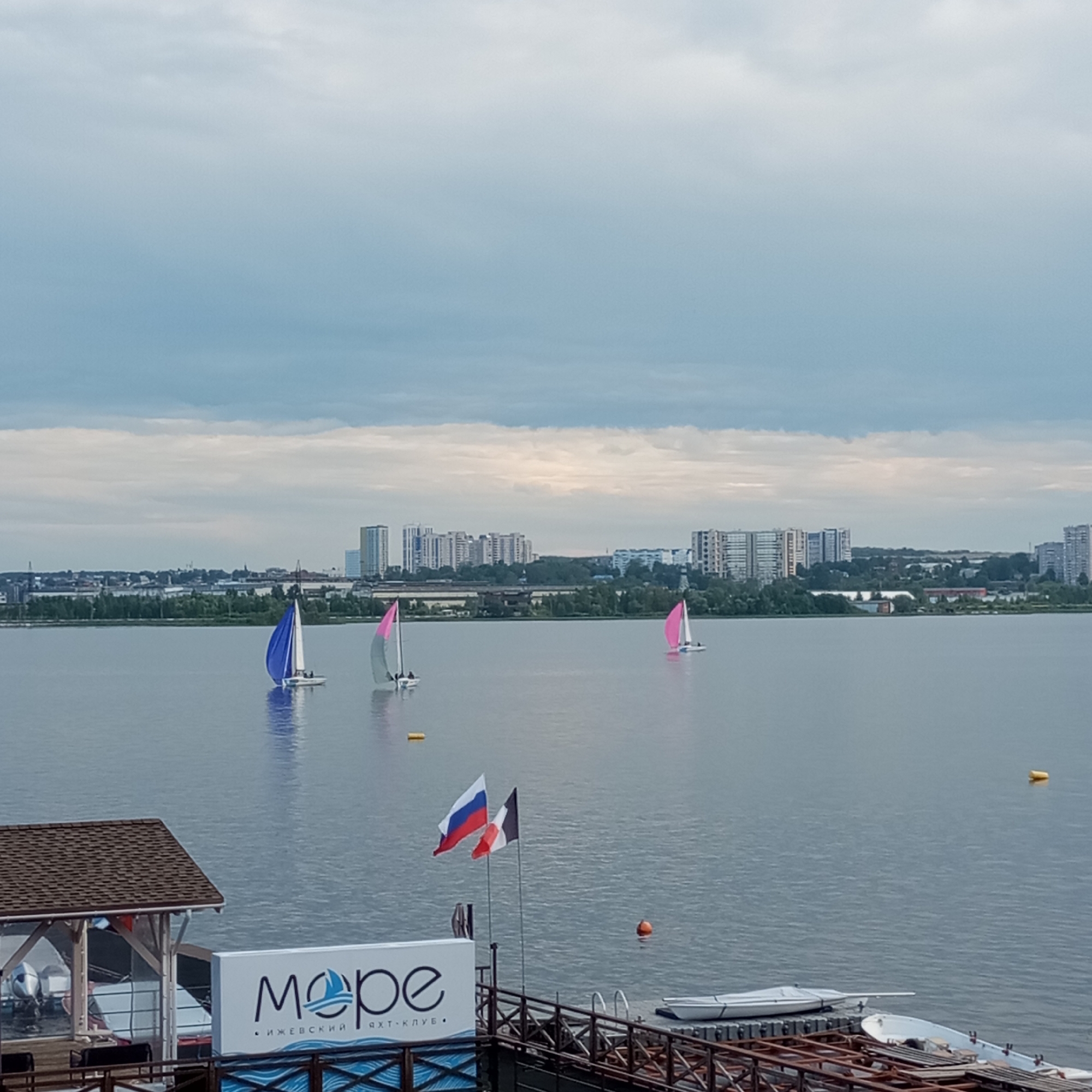 The image depicts a wide view of the Izhevsk Pond in Izhevsk, Russia, on a cloudy day. In the foreground, there is a wooden pier with a building structure to the left, featuring a brown tiled roof and outdoor seating. A white sign with the word "море" (meaning "sea" in Russian) and a stylized sailboat logo is prominently displayed on the pier. Two flags, the Russian flag and a red, white, and blue tricolor flag (possibly French), are planted on poles on the pier.
In the middle ground, several sailboats are visible on the water. One sailboat on the left has a bright blue sail, and two others to the right have pink sails. The water is calm, reflecting the overcast sky.
The background shows the Izhevsk skyline, characterized by a mix of residential buildings and industrial structures. Trees line the shore between the water and the city. The overall scene is one of a serene, urban waterfront environment.