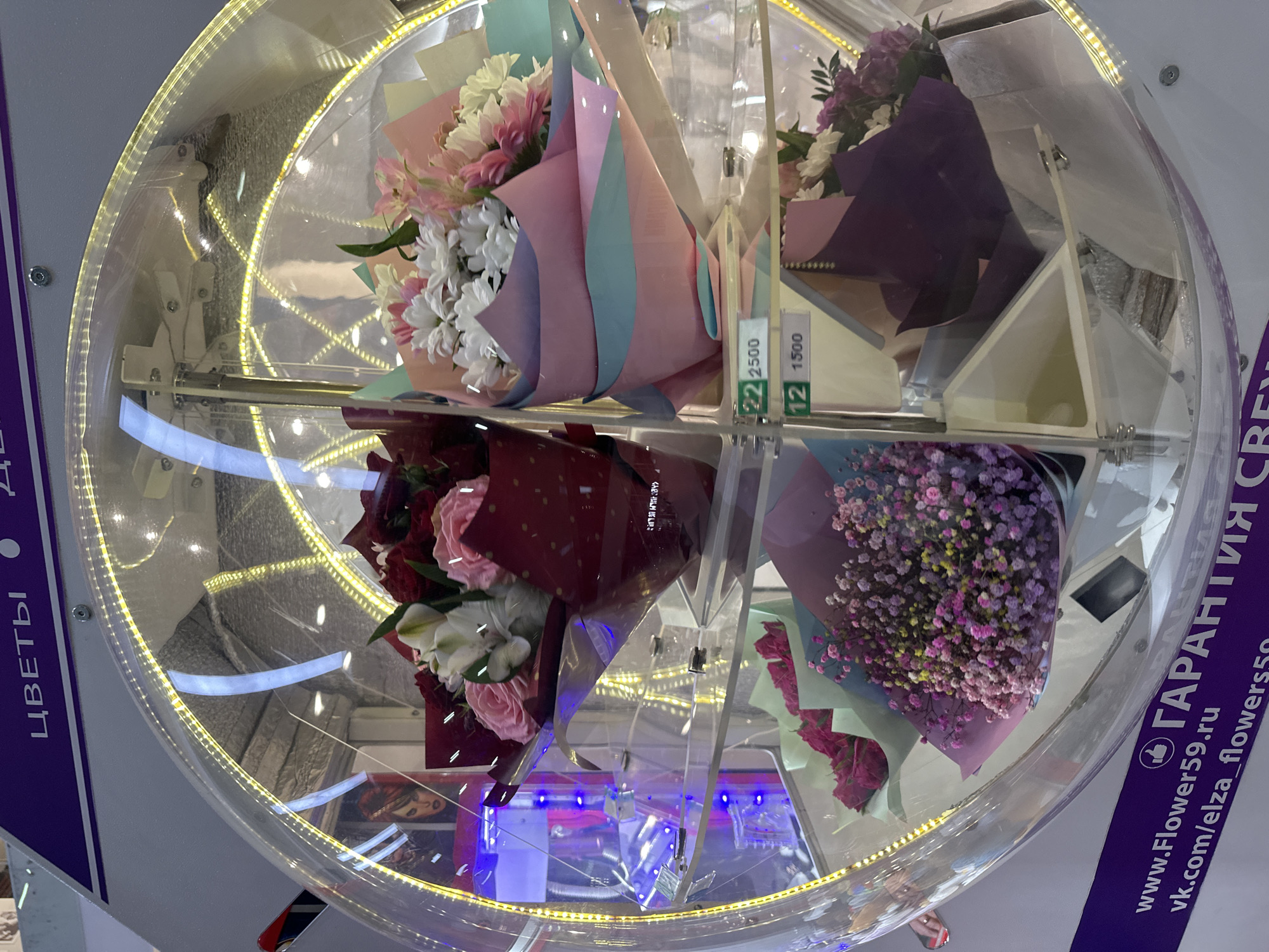 The image shows the inside of a flower vending machine. There are four bouquets of flowers: one containing pink and white flowers wrapped in pink and blue paper, another containing purple and white flowers in purple paper, a third containing red and white roses in maroon paper, and a final bouquet of small pink and purple flowers wrapped in matching colored paper. The machine is made of clear plastic or glass and is illuminated with a yellow light. A sign on the side includes a web address indicating this is a flower vending machine from elza_flowers59.ru in Bolshoye Savino, Russia.