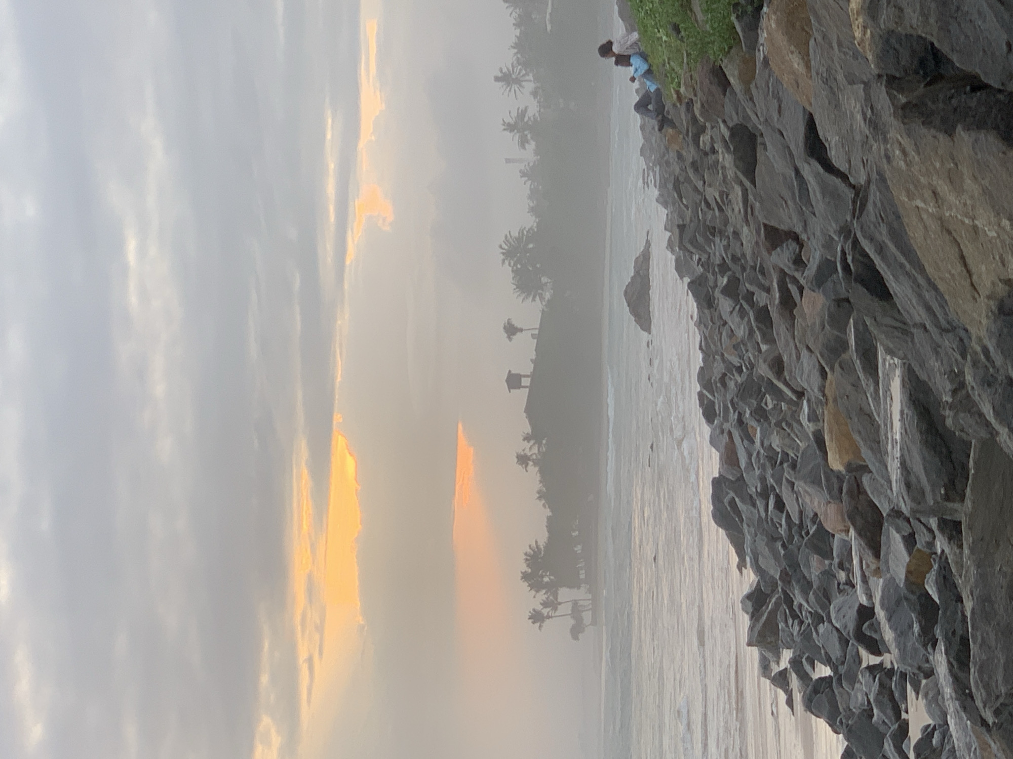 The image shows a coastal scene in Kathaluwa, Sri Lanka. The sky is overcast with diffused sunlight, possibly during sunset. Palm trees line the horizon amidst a misty atmosphere. A rocky embankment forms the right edge of the image, sloping down towards the waterline. A person sits on the embankment, their features indistinct due to distance and atmospheric haze. The sea is calm, with subtle wave action visible.
