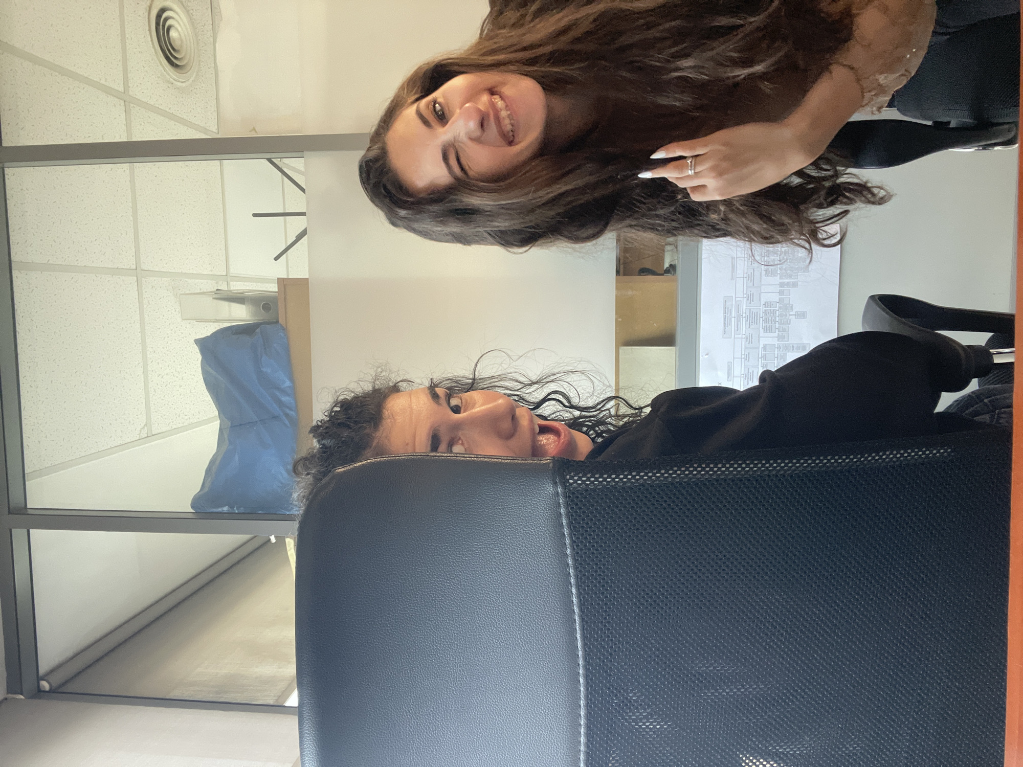 Two young women are captured in a playful indoor office setting. The image, originally rotated, shows them appearing from the top and bottom of the frame.
The woman at the top has long, wavy dark brown hair and is smiling widely at the camera, her eyes crinkling. She wears a ring on her left hand's ring finger, and her fingernails are painted white. Her expression conveys happiness and amusement.
The second woman, positioned in the lower part of the frame, has dark, curly hair and is peeking over the back of a dark office chair, which is prominently featured in the foreground. Her mouth is open in a surprised or playful expression. The chair appears to have a mesh backrest.
The background reveals typical office elements: a white acoustic tile ceiling with a circular air vent, glass partitions separating different areas, and a partially visible document or chart hanging on a wall, too blurry to read. A blue bag or object is visible behind one of the glass panels. The overall atmosphere is lighthearted, suggesting a casual moment between colleagues or friends in an office in Sofia, Bulgaria.