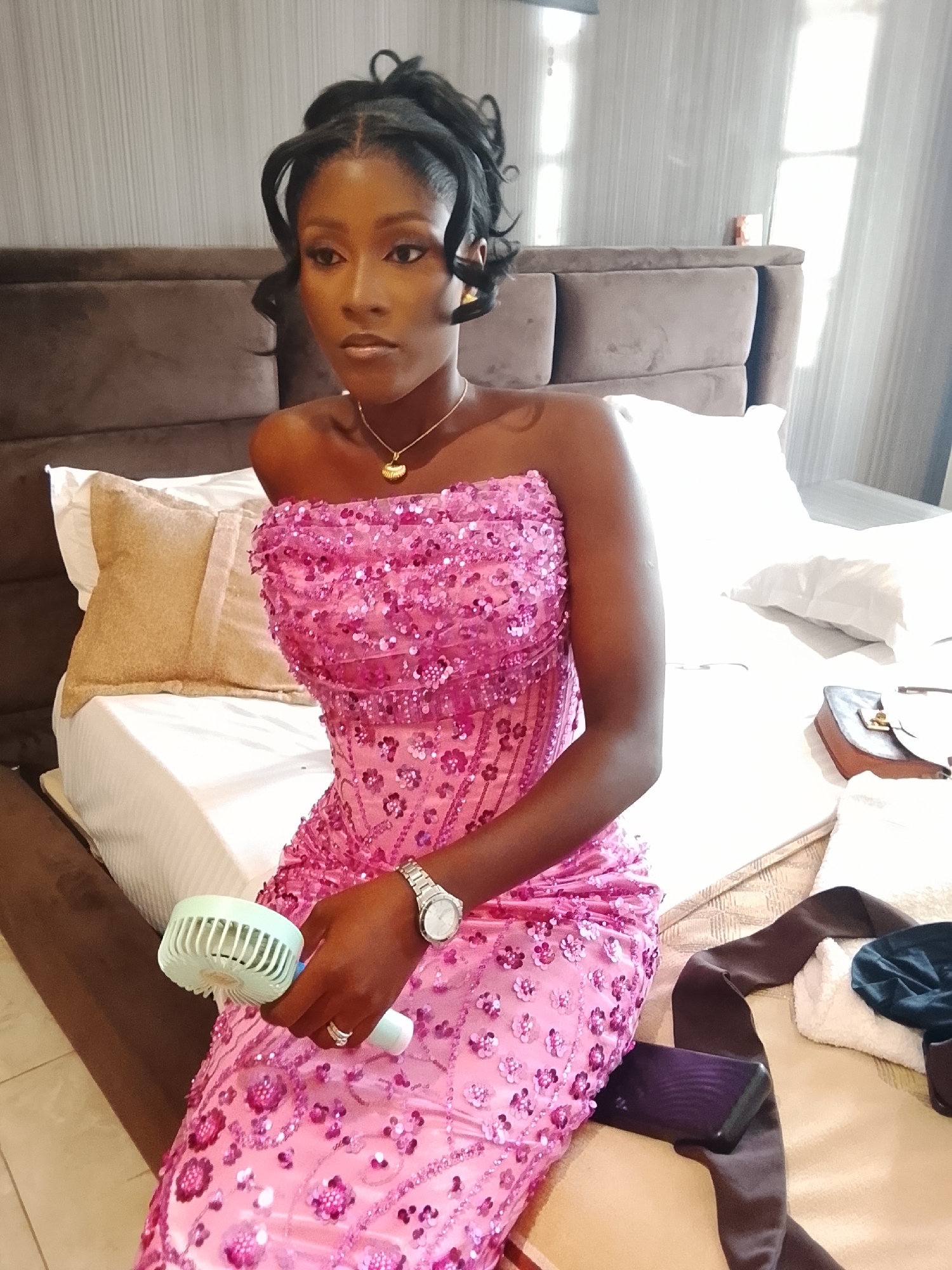 A medium shot depicts a dark-skinned woman seated on a bed, presumed to be indoors. She wears a pink, strapless, sequined dress. The woman is also wearing a gold necklace, a watch on her left wrist, and rings. She holds a small, handheld fan in her right hand. Her hair is styled up.
Behind her is a bed with brown headboards, white linens, and decorative pillows. A purse, a mobile phone, a brown sash, a white towel, and a dark-colored fabric are also on the bed.
The location is inferred to be in Gwarinpa, Nigeria, as specified in the prompt's instructions.