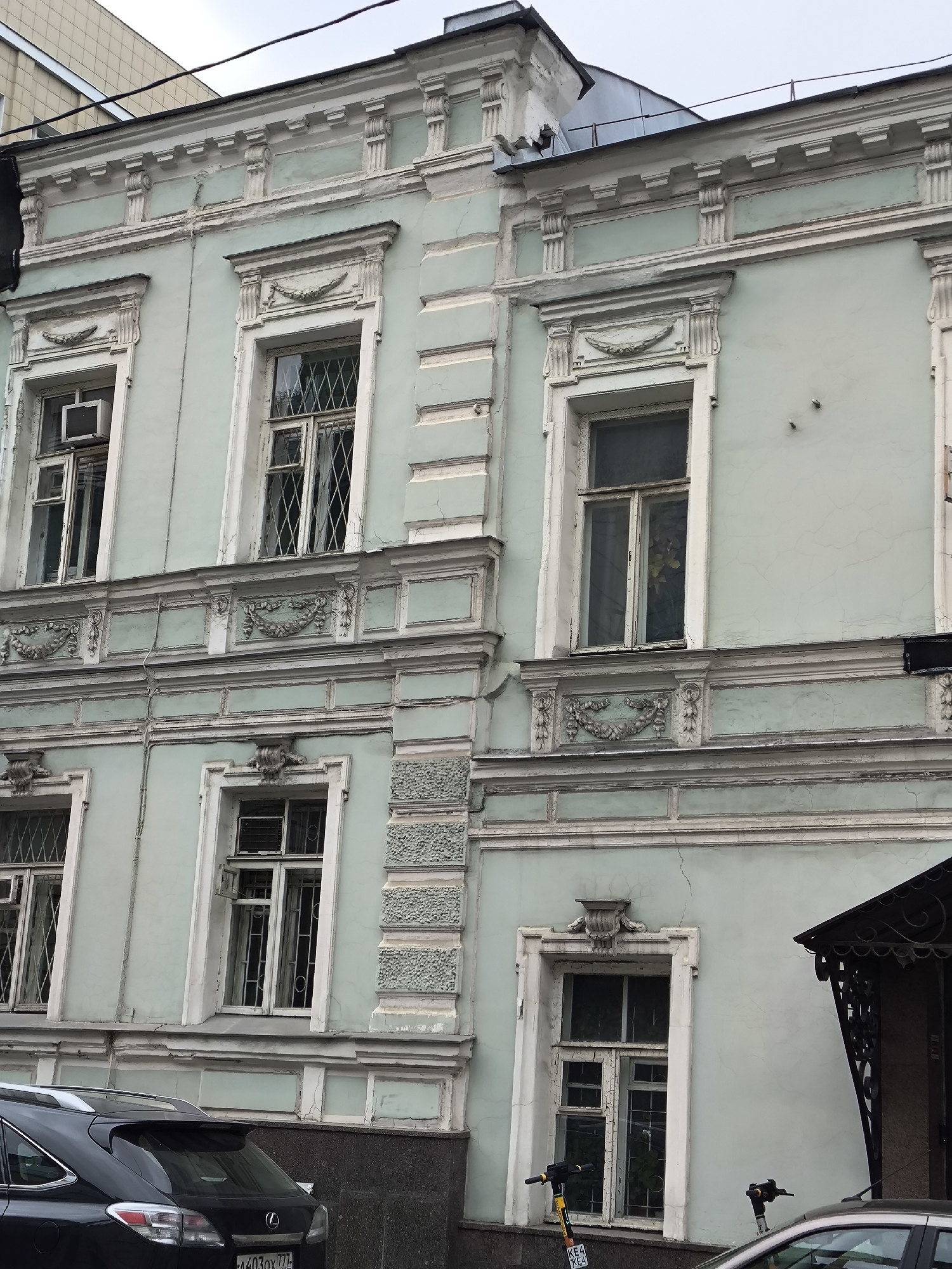 The image depicts a pale green, two-story building facade in Moscow, Russia. The architectural style is characterized by ornate window frames and decorative moldings. The windows are tall and narrow, some with lattice-like grilles. An air conditioner unit is visible in one window. The building's lower level has a dark stone base. Parked cars are visible in the foreground, along with two electric scooters standing on the sidewalk. The sky is overcast.