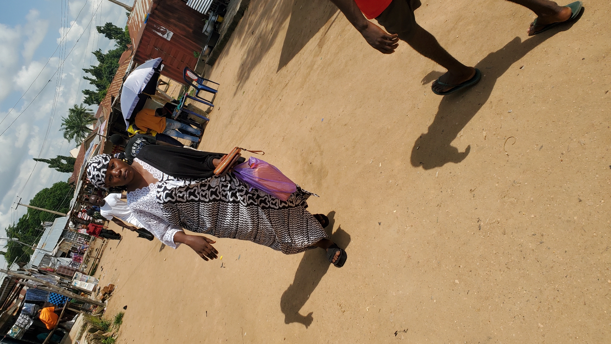 The image depicts a street scene in Jalingo, Nigeria, under a partly cloudy sky. The foreground features the lower body of a person walking, casting a distinct shadow on the sandy ground. Further into the scene, a woman is captured in motion, wearing a patterned headscarf, a black and white patterned dress, and black sandals. She is holding a light purple plastic bag and a brown wallet. Behind her, other individuals and market stalls line the dusty street, with corrugated metal roofs and various goods visible. A white and blue umbrella stands near a stall, and several blue plastic chairs are set up. The environment suggests a bustling marketplace with temporary structures.