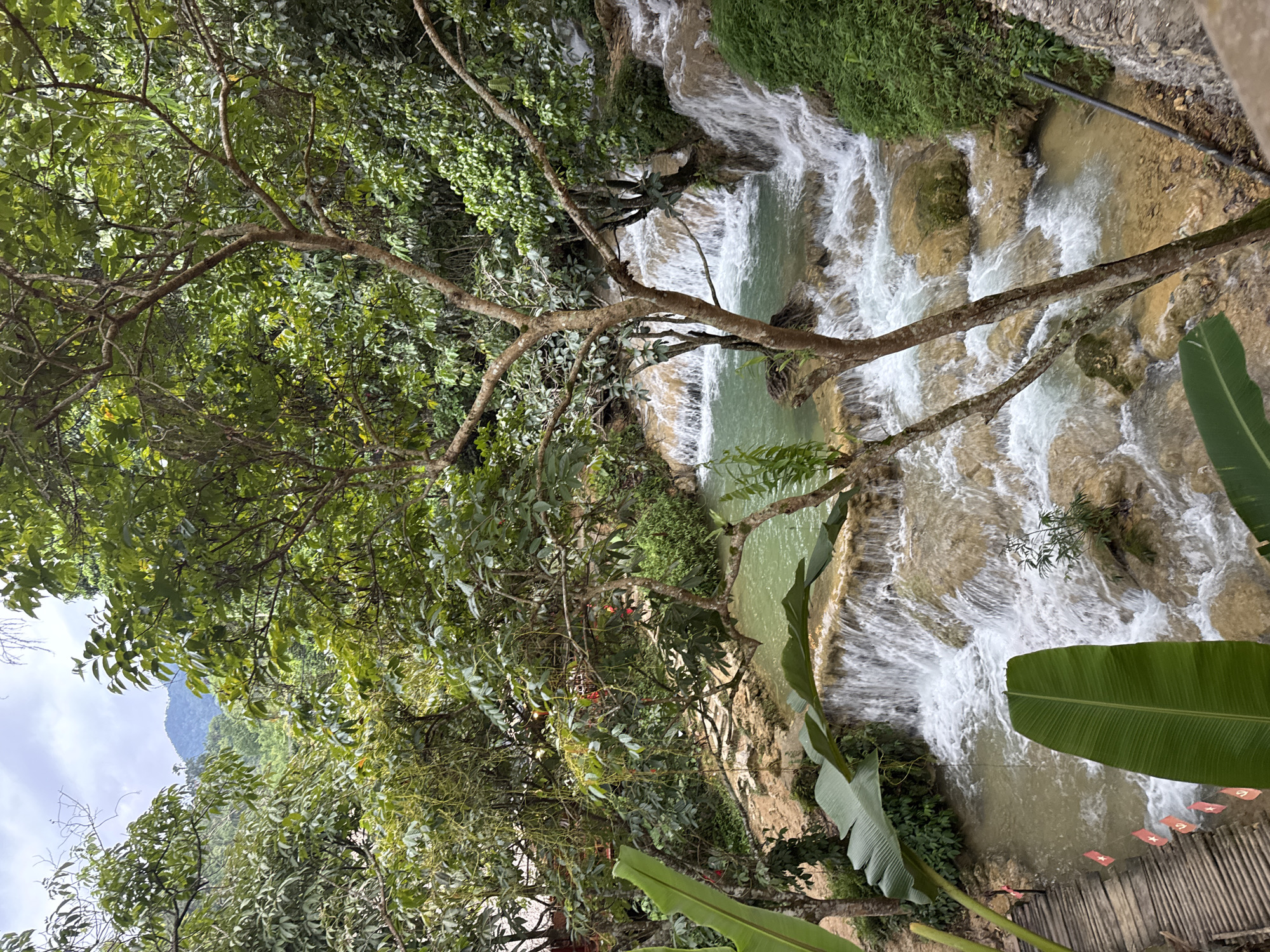 The image depicts a natural landscape featuring a multi-tiered waterfall with clear, turquoise water flowing over light brown rocks and cascading into pools. Lush green foliage, including large banana leaves and dense trees with visible branches and smaller leaves, surrounds the waterfall. A wooden walkway, made of planks and elevated slightly, is visible on the left side of the image, adorned with small red flags. In the background, a distant mountain covered in vegetation can be seen under a cloudy sky. The scene is evocative of a tropical or subtropical environment, likely a natural park or scenic area. The presence of the walkway suggests it is an accessible tourist destination within the Cổ Lũng Commune, Vietnam, likely in a rural or mountainous region, as indicated by the natural terrain and vegetation.