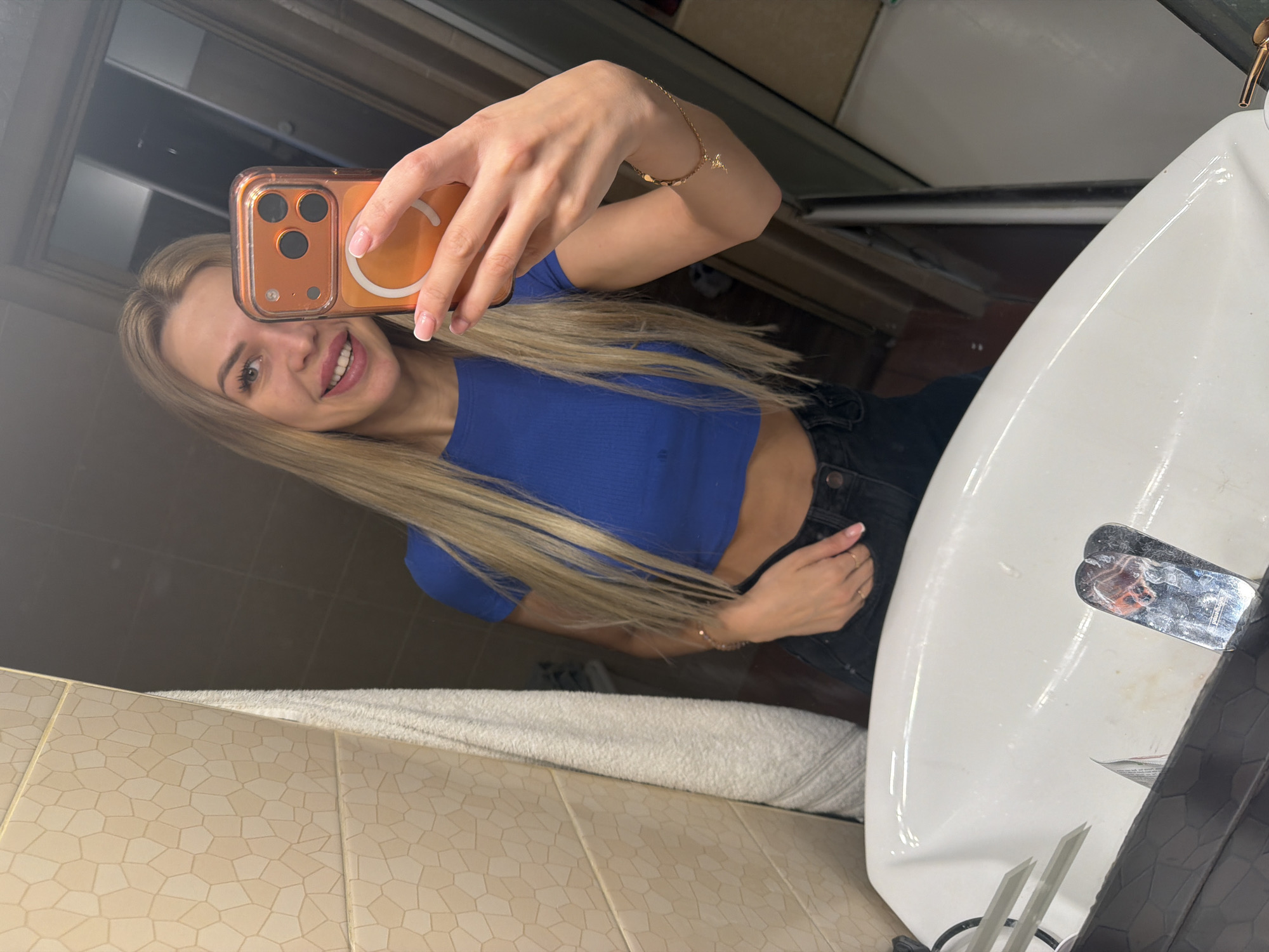 A blonde woman in a blue cropped t-shirt and black jeans is taking a mirror selfie in a bathroom. She is holding an orange phone with a clear case. The bathroom has beige tiled walls with a textured pattern and a white sink. A gray towel hangs on the wall to the left of the mirror. The woman is smiling and her hair is long and straight. She is wearing rings on her fingers and a bracelet on her wrist. The scene is captured from a medium shot, showing the woman from her chest up and the sink in the foreground. The reflection in the mirror shows the woman and the bathroom environment.