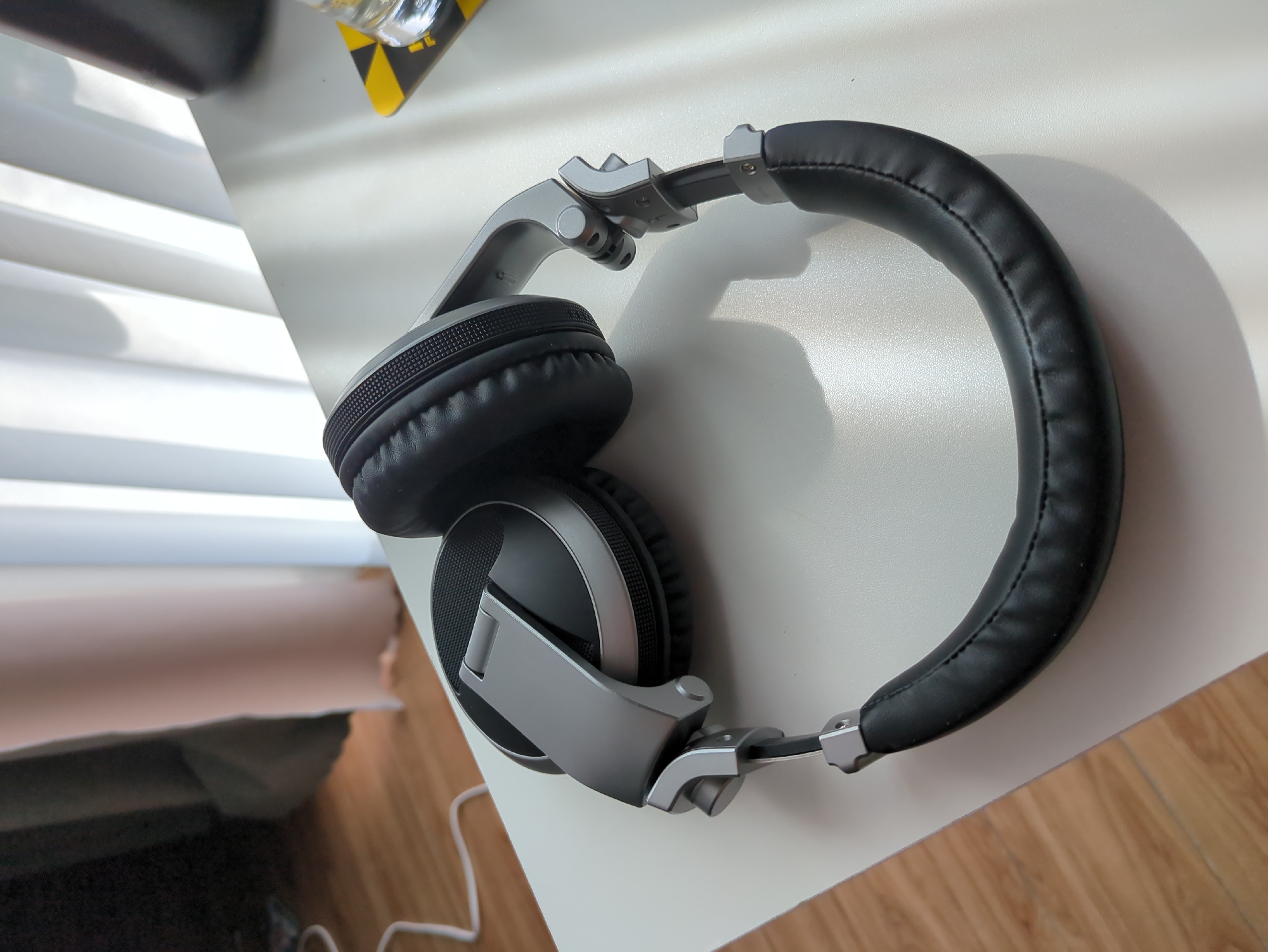 A pair of black over-ear headphones rests on a white surface, likely a desk. The headphones are silver and black, with plush black earcups and a padded headband. They are positioned to show the side of one earcup and the curve of the headband. The white surface is clean and appears to be a flat tabletop. In the background, vertical blinds are visible, casting shadows on the surface, suggesting a window or light source nearby. To the right of the headphones, a coaster with a yellow and black hazard pattern and a glass are partially visible. To the left, a white cable trails away from the headphones. The overall scene suggests a home or office environment, possibly in Đà Nẵng, Vietnam, given the ambient light and implied setting.