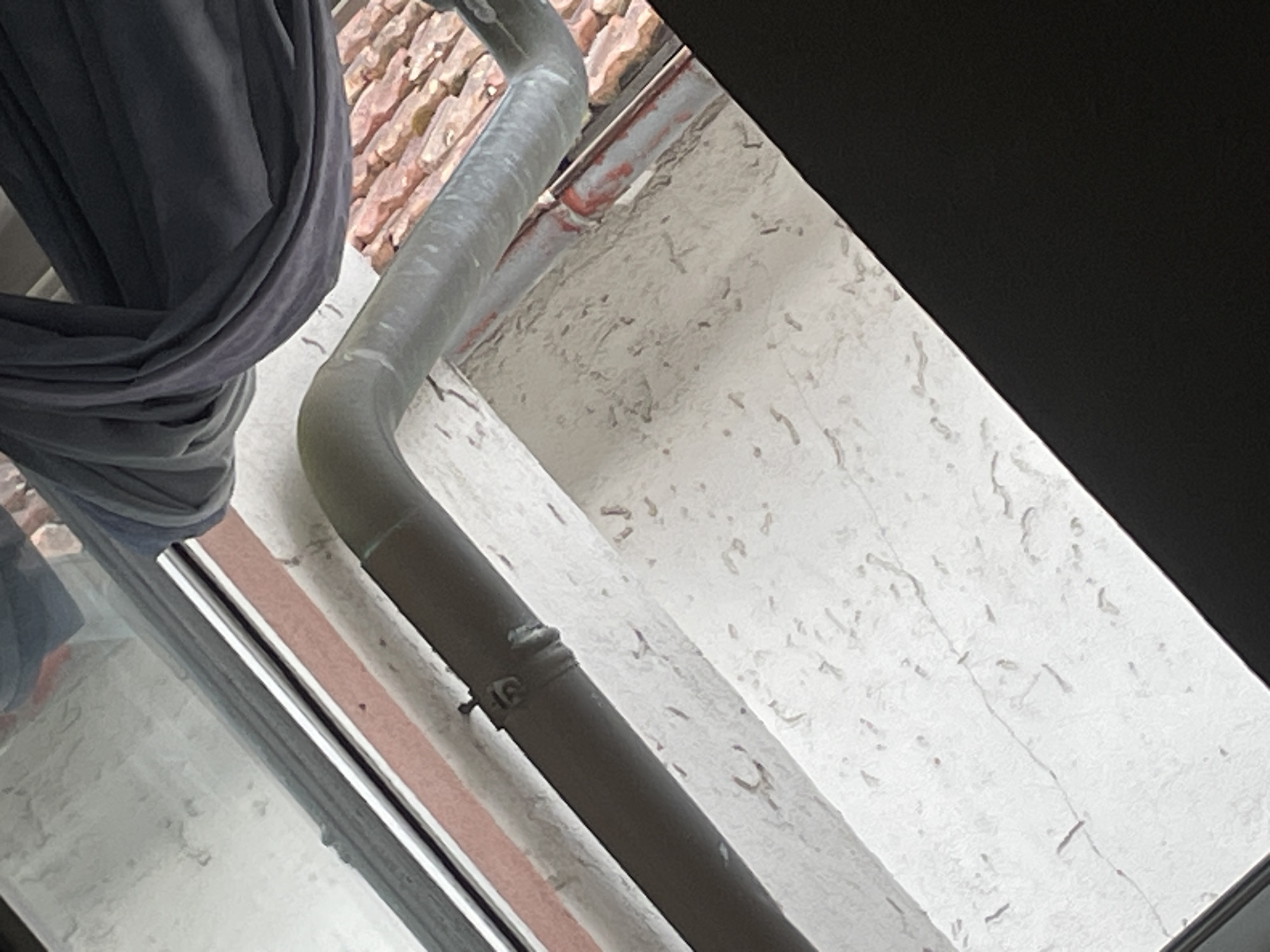 A close-up, angled shot looks down from a window onto a textured, off-white exterior wall. A dark gray downspout runs diagonally across the frame, secured to the wall by a metal clamp. To the right of the downspout, a section of reddish-brown roof tiles is visible, suggesting the structure is likely a house or other building. A dark gray fabric, possibly a curtain or drape, is bunched up and hangs loosely near the top of the frame, partially obscuring the view of the sky and roof. The lower right of the image is dominated by a large, solid black area, which could be a portion of the window frame or another obstruction. The overall impression is of an outdoor urban environment, possibly a residential area in Hohberg, Germany, given the details like the downspout, wall texture, and roof tiles. No people or clear actions are depicted.