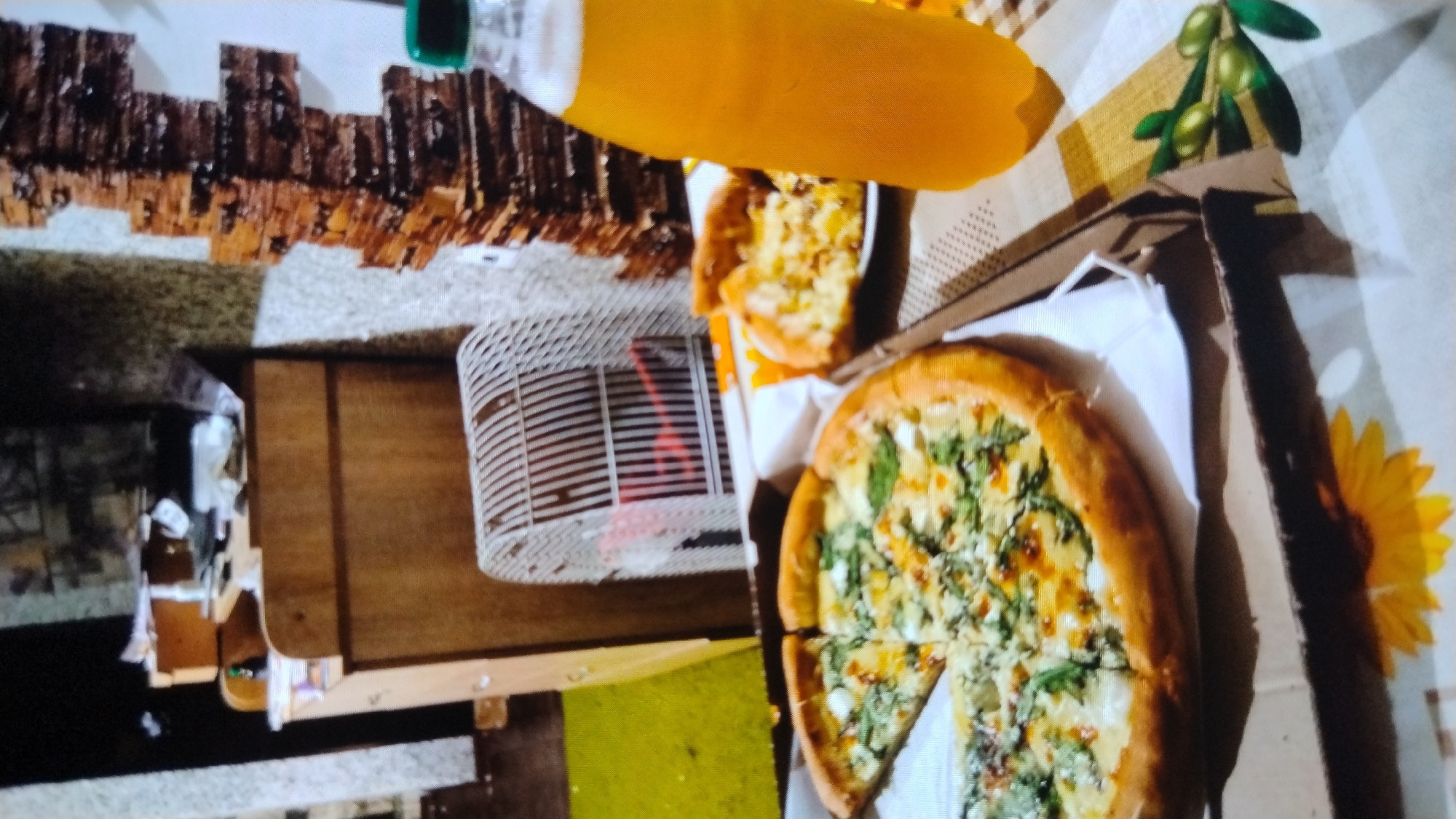 The image depicts a meal and a drink on a table, with a blurred background suggesting an indoor domestic setting. In the foreground, a partially eaten pizza is visible in a cardboard box. To the right, a plastic bottle of yellow liquid, likely a beverage, stands upright. Behind the pizza and drink, a white wire cage, possibly for a pet, is visible, and further back, a wooden cabinet is present. The overall ambiance is casual and informal. The location is stated to be the city of Mykolaiv, Ukraine.