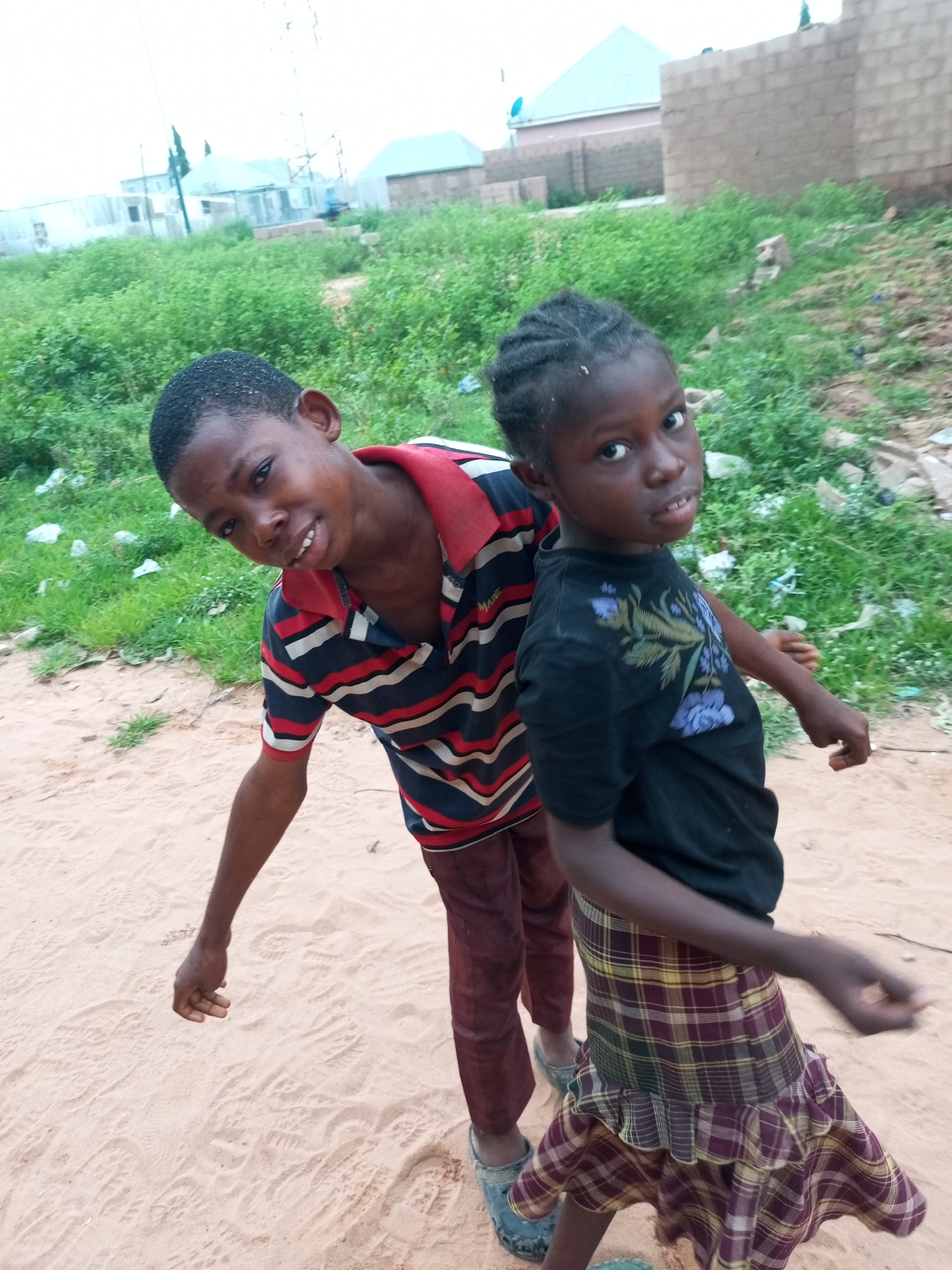 In Tempure, Nigeria, two young children are captured in a candid moment outdoors. The child on the left, a boy, is looking towards the camera with a slight smile. He wears a horizontally striped shirt in red, navy, and white, with a red collar. His pants are a dark reddish-brown. The child on the right, a girl, faces the camera directly, her expression more serious. She has braided hair and wears a black t-shirt with a floral design in shades of blue and green. Her skirt is a plaid pattern in shades of maroon, yellow, and green. They are standing on a sandy ground, with patches of green vegetation and unfinished brick buildings visible in the background, suggesting a developing urban or semi-urban environment. The lighting suggests it is daytime, possibly overcast.