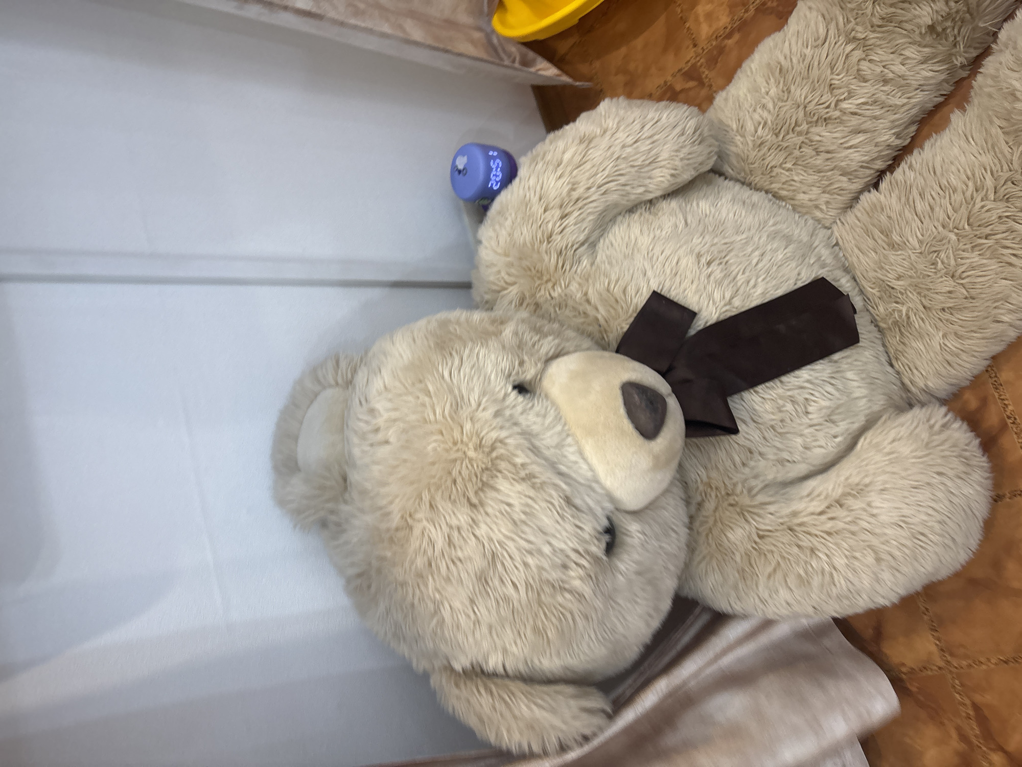 A beige teddy bear with a brown bow tie rests on a white surface alongside a blue-topped bottle. The teddy bear appears fluffy, and the brown bow is securely fastened around its neck. Part of a brown, textured surface is visible in the top right corner, potentially indicating a wooden surface or rug. The background lacks distinct features and specific details to establish the location as Ферма, Russia, or any other specific place.