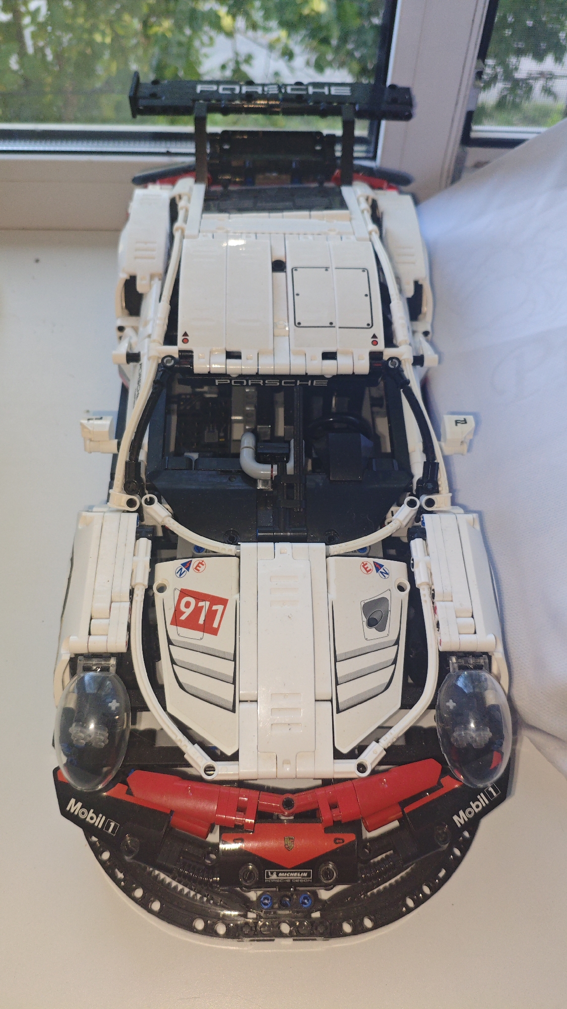 A LEGO Technic Porsche 911 RSR model car is displayed on a white surface, possibly a windowsill, with a blurred green foliage background suggesting an outdoor environment. The car is white with black and red accents and features various sponsor decals, including "PORSCHE," "911," "Mobil 1," and "MICHELIN." The model is highly detailed, showcasing elements like the spoiler, headlights, and interior cockpit. There are no people or animals visible in the image. The lighting suggests it is daytime, and the overall scene is static and still. The location of Serebriansk, Kazakhstan is not visually represented in the image.