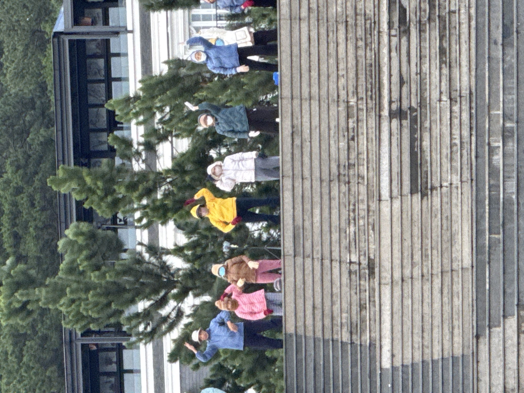 In Da Lat, Vietnam, a group of eight individuals are positioned on a wide, tiered concrete staircase. The people are diverse in their attire, wearing various colors of jackets, pants, and headwear. Some are wearing face masks and gloves. All are facing towards the viewer and appear to be waving. Behind them, lush green pine trees and a modern building with a dark roofline are visible. The scene conveys a sense of activity and engagement.