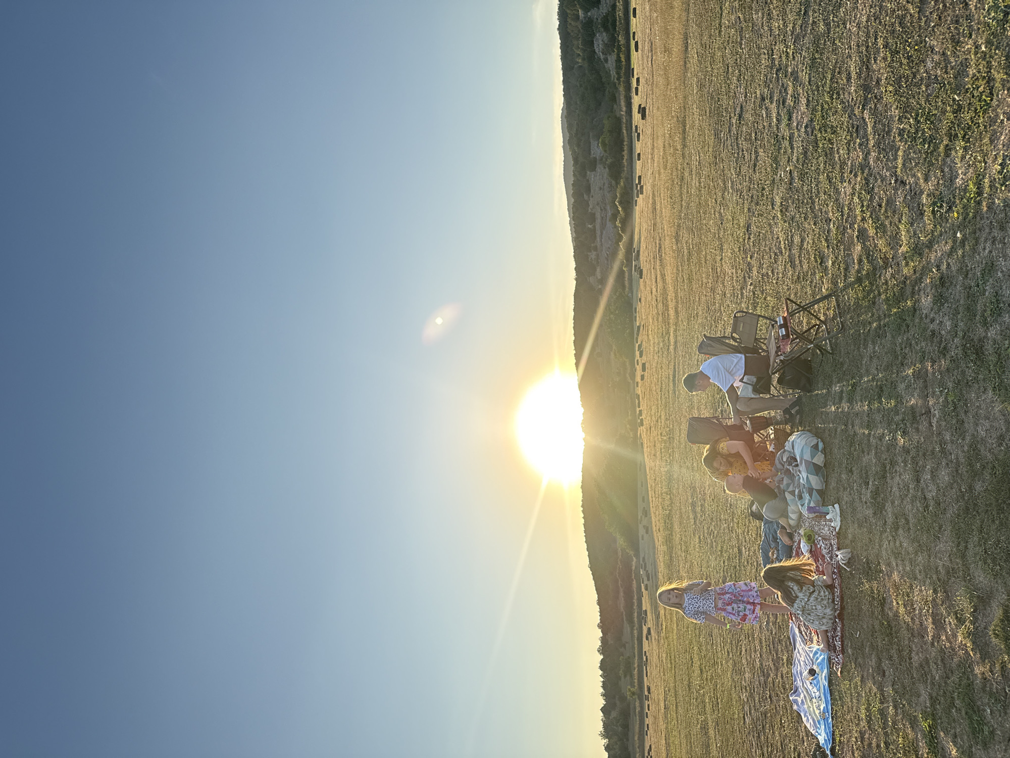 The image depicts a family picnic at sunset in a grassy field with rolling hills in the background. A family, consisting of at least two adults and three children, is gathered on blankets and camping chairs. The sun is low on the horizon, casting a warm glow and long shadows across the scene. Several hay bales are scattered in the field. The location appears to be in the Орлиновский округ, Ukraine, given the context provided.