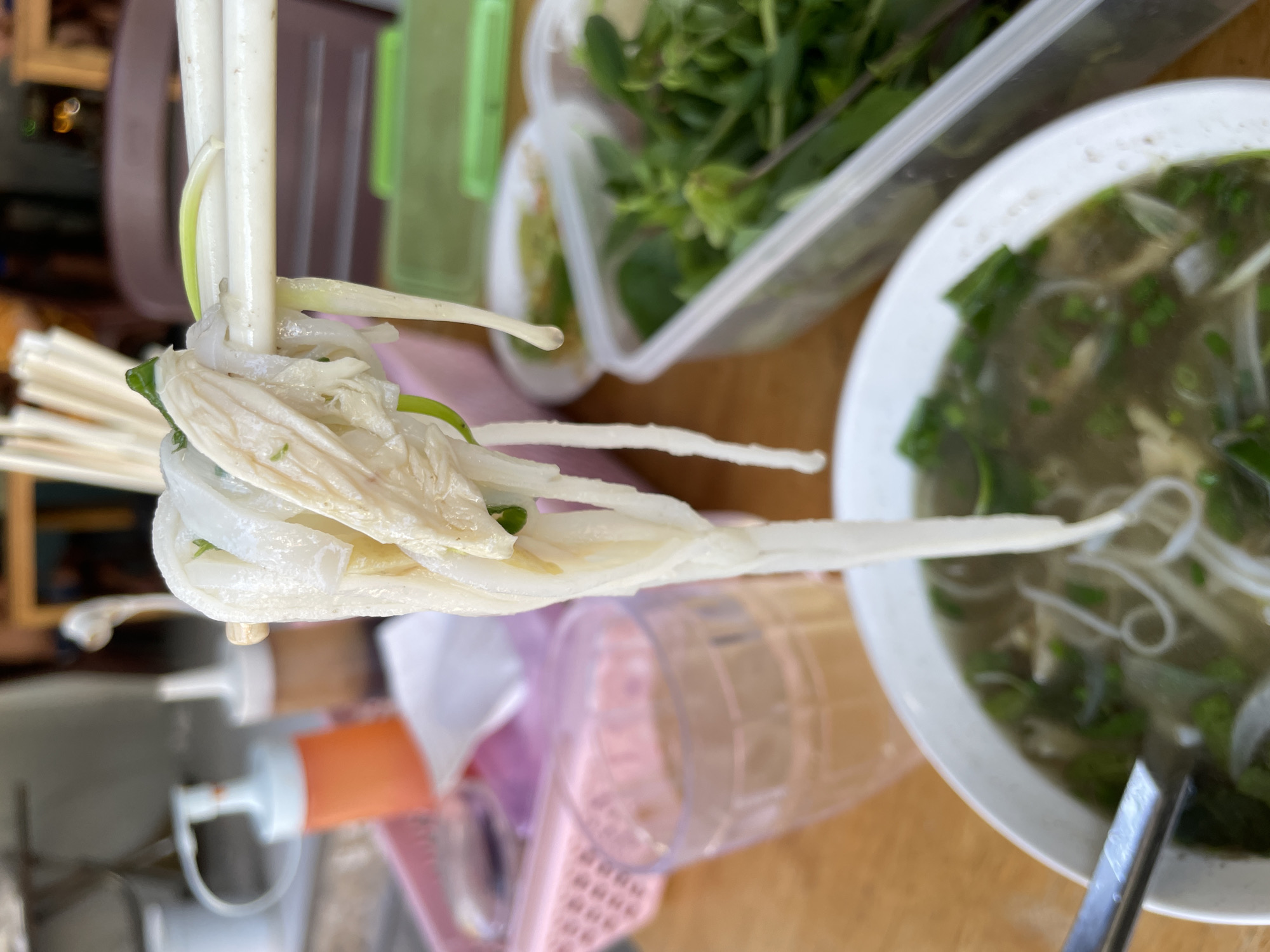 The image depicts a close-up view of a bowl of pho, a Vietnamese noodle soup, at a food stall in Phu Thuy Ward, Vietnam. Chopsticks are holding a portion of the noodles and chicken above the bowl, with a droplet of soup about to fall. The background shows condiments, a glass of liquid, and a container of fresh herbs, typical of a Vietnamese eatery. The lighting suggests an outdoor setting during the daytime.