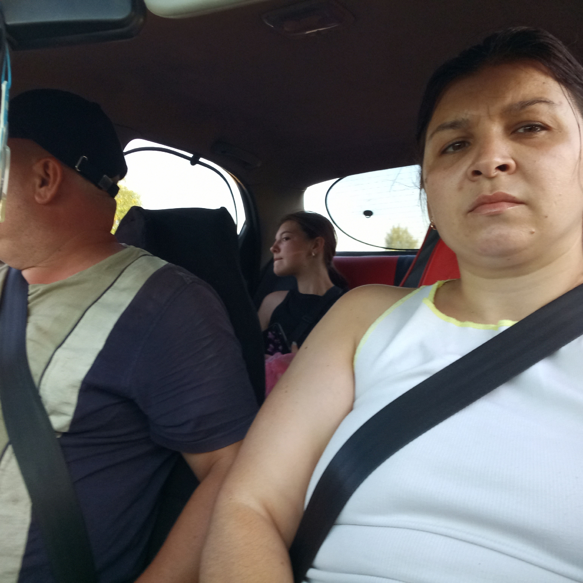 The image is a selfie taken from inside a car in Kropyvnytskyi, Ukraine. The foreground is dominated by the faces of two women and the shoulder of a man. The woman on the right is closer to the camera, with her face partially visible and her arm on the right side. She is wearing a white top with a yellow trim and a black seatbelt. To her left, a man wearing a dark blue shirt with light-colored stripes and a black cap is visible from the side, also with a seatbelt. In the background, a younger woman with dark hair is seated in the back of the car, looking to the left. She is wearing a black top. The car's interior is visible, including the headrests and parts of the windows. The lighting suggests it is daytime.