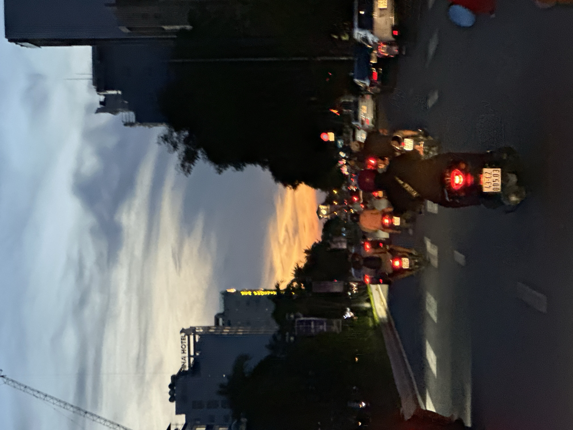 The image depicts a street scene in Da Nang, Vietnam at dusk. A crane is visible in the upper left corner, adjacent to a building with the text "ANA HOTEL" on its facade. A busy road is in the foreground, with numerous motorbikes and a few cars navigating the traffic. The sky is a gradient of blue and orange, indicating the sunset. The overall atmosphere is one of urban activity during twilight.