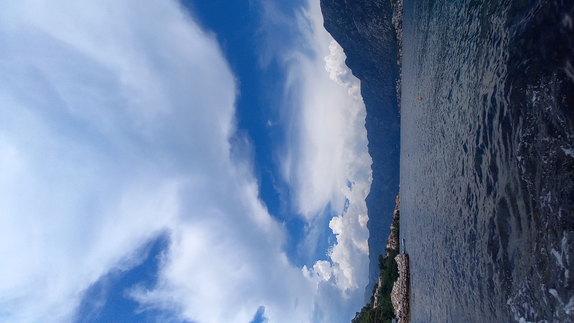 The image depicts a serene seascape with a view of the Bay of Kotor, likely from the vicinity of Muo, Montenegro. The foreground is dominated by the dark, rippling surface of the water, with white foam and ripples indicating gentle movement. In the mid-ground, a calm expanse of the bay stretches towards a distant, mountainous coastline.
Along the shoreline, buildings of various sizes and colors are visible, suggesting a populated area nestled at the foot of the mountains. Some structures appear to be residential, with tiled roofs and multi-story designs. To the left, a rocky embankment with some vegetation juts into the water, possibly a man-made fortification or a natural rocky outcrop.
The background is characterized by a range of dark, imposing mountains that rise steeply from the water's edge. Their slopes are covered in dense vegetation, and their peaks are shrouded in a slightly hazy atmosphere, suggesting they are quite distant.
The sky above is a dramatic display of clouds. Large, white, cumulus clouds are interspersed with patches of deep blue sky. The clouds are textured and voluminous, with some areas appearing dense and others more wispy, hinting at dynamic weather conditions. The overall lighting suggests it is daytime, with diffused sunlight filtering through the clouds, casting a soft glow on the scene. No people or animals are clearly visible in the image.