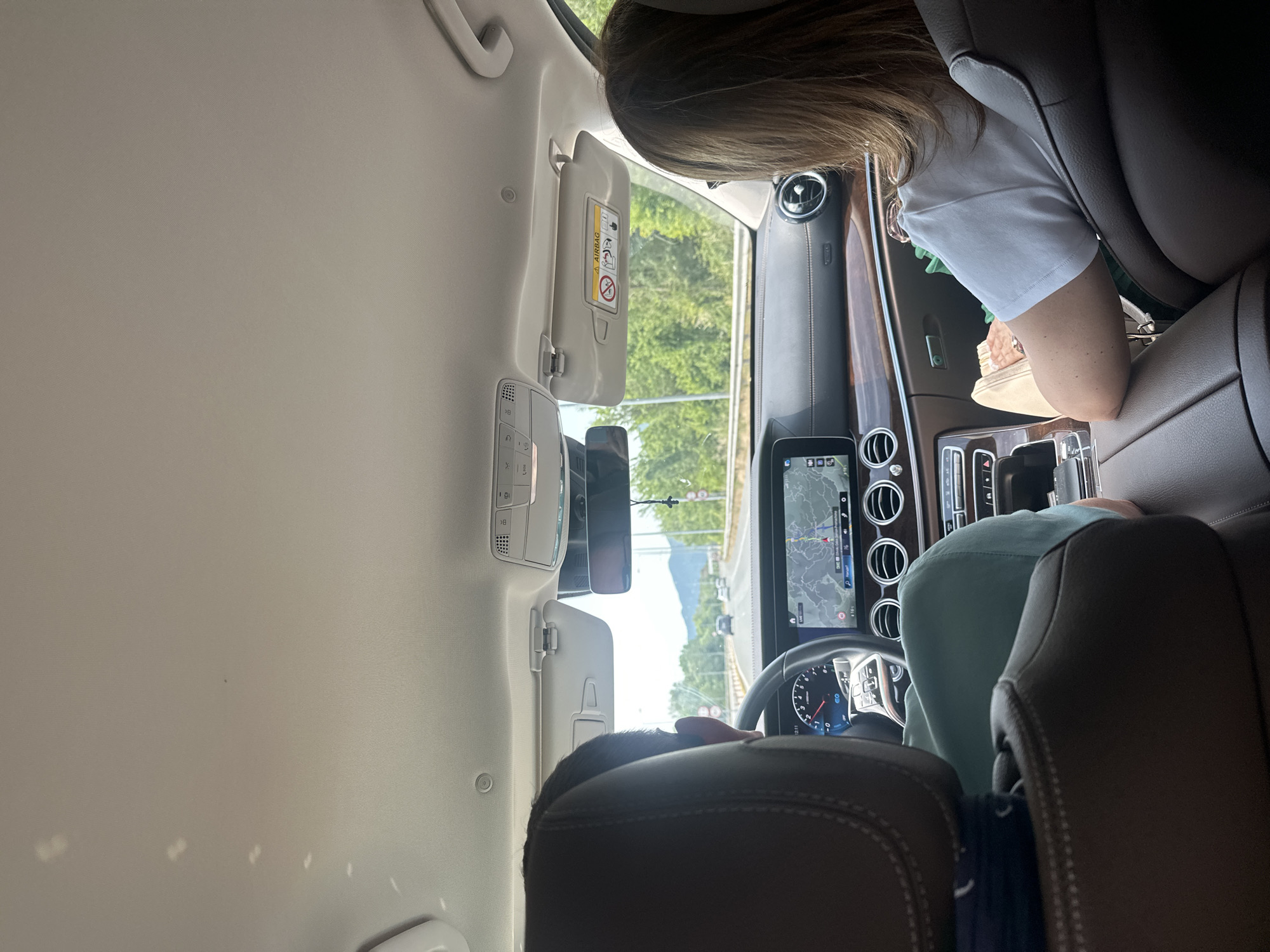 The image depicts the interior of a modern car from the perspective of a rear passenger seat, looking towards the front. The setting is daytime, with bright natural light suggesting clear, sunny weather.
Two people are present in the car. In the front passenger seat, a person with light brown hair, wearing a white t-shirt, is partially visible, seemingly holding a small, light-colored bag or wallet in their lap. The driver's head and shoulder, wearing a light blue or green shirt, are also visible, situated behind the steering wheel.
The car's interior features a sophisticated dashboard with multiple circular air vents and a large central display screen showing a navigation map. The time "12:11" is visible on the screen, and the dashboard also shows "km/h" and possibly "EQ," hinting at a Mercedes-Benz vehicle. A dashcam is attached to the rearview mirror. The passenger-side sun visor is lowered, displaying a yellow "A AIRBAG" warning label with universal safety pictograms, including warnings against placing a rear-facing child seat in the front.
Through the windshield, a scenic view unfolds, with lush green trees lining the road and prominent mountains in the distance under a bright sky. This landscape is consistent with the provided location context of Δημοτική Ενότητα Προμαχώνος, Greece, suggesting a journey through a picturesque, possibly mountainous region. The overall scene conveys a sense of a road trip or leisurely drive.