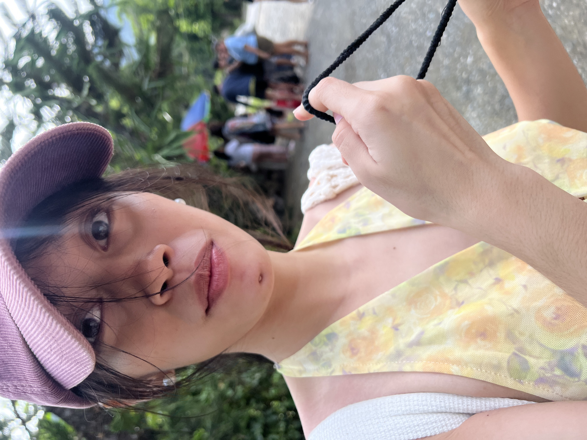 A young woman poses for a selfie. She is wearing a pink corduroy cap and a yellow floral patterned top with a white crocheted collar. She holds a black cord in her right hand. In the blurry background, several people are visible. The location is Banaybanay, Philippines.