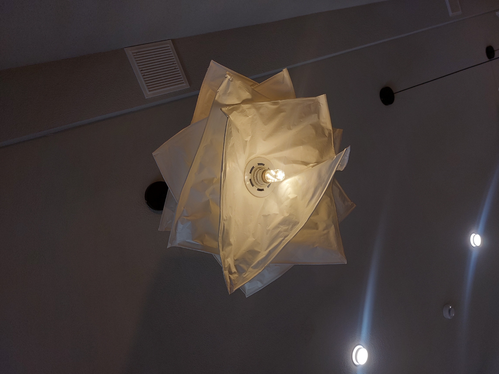The image shows a close-up, low-angle shot of a unique, multi-layered white paper lampshade hanging from a dark ceiling. A bright incandescent lightbulb is visible in the center of the lampshade, illuminating the translucent material from within. Several smaller, circular lights are embedded in the ceiling in the background, along with a rectangular vent. The ceiling is a dark gray, textured material, and the overall lighting is dim, with the lampshade being the primary light source. There are no people or animals present in the image. The location is not specified in the image, but it is described as being in the city of Kyzylorda, Kazakhstan.