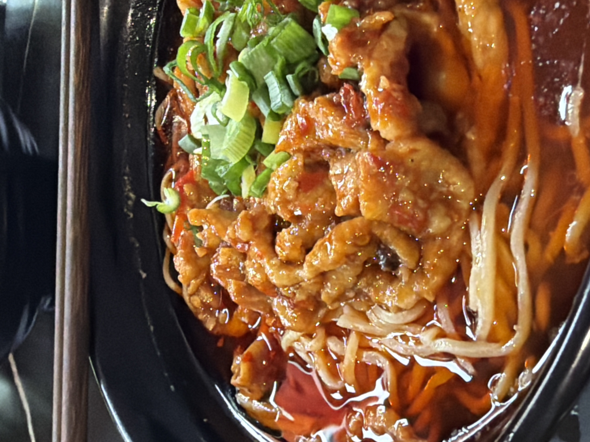 A close-up, high-angle shot shows a black bowl filled with what appears to be a noodle dish. The dish consists of noodles, what may be caramelized meat, and chopped green onions. The noodles and meat are coated in a reddish-brown sauce. Chopsticks are visible to the left of the bowl. The location is unknown, though Saint Petersburg, Russia, was specified in the prompt.