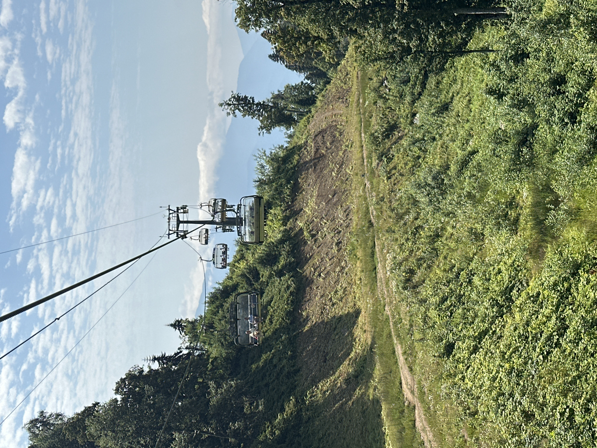 The image depicts a ski lift ascending a lush, green mountainside under a bright blue sky with scattered white clouds. The ski lift, with multiple open-air cabins, is the central focus of the composition. In one of the cabins, two individuals are visible, seated and facing forward, likely enjoying the scenic ride. The mountainside is covered in dense vegetation, with a winding dirt path visible on the left side of the frame. The background reveals layers of hazy blue mountains, suggesting a vast, natural landscape. The scene is set in Estosadok, Russia, a location known for its mountainous terrain and ski resorts. The overall impression is one of natural beauty and outdoor recreation.