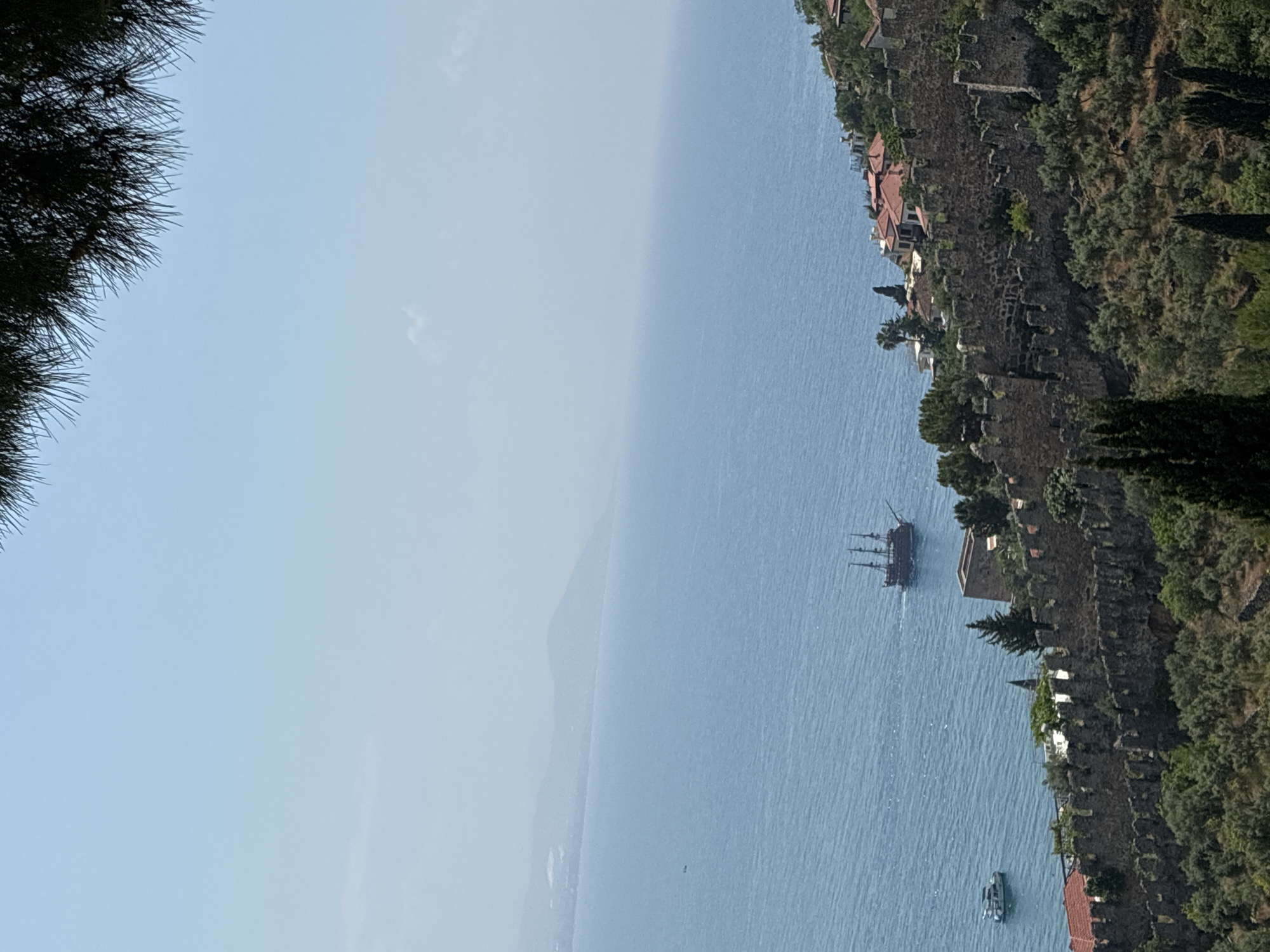 The image depicts a scenic view of Alanya, Turkey, with the Alanya Castle walls prominently featured in the foreground. The ancient stone fortification stretches across a verdant hillside, overlooking the azure waters of the Mediterranean Sea. A tall, traditional sailing ship with multiple masts is visible on the sea, moving away from the viewer. Near the shore, a modern white boat is also present. The sea extends to the horizon, where it meets a hazy mountain range. The sky above is a clear, light blue with a few wispy clouds. The overall impression is one of historical grandeur and coastal beauty.