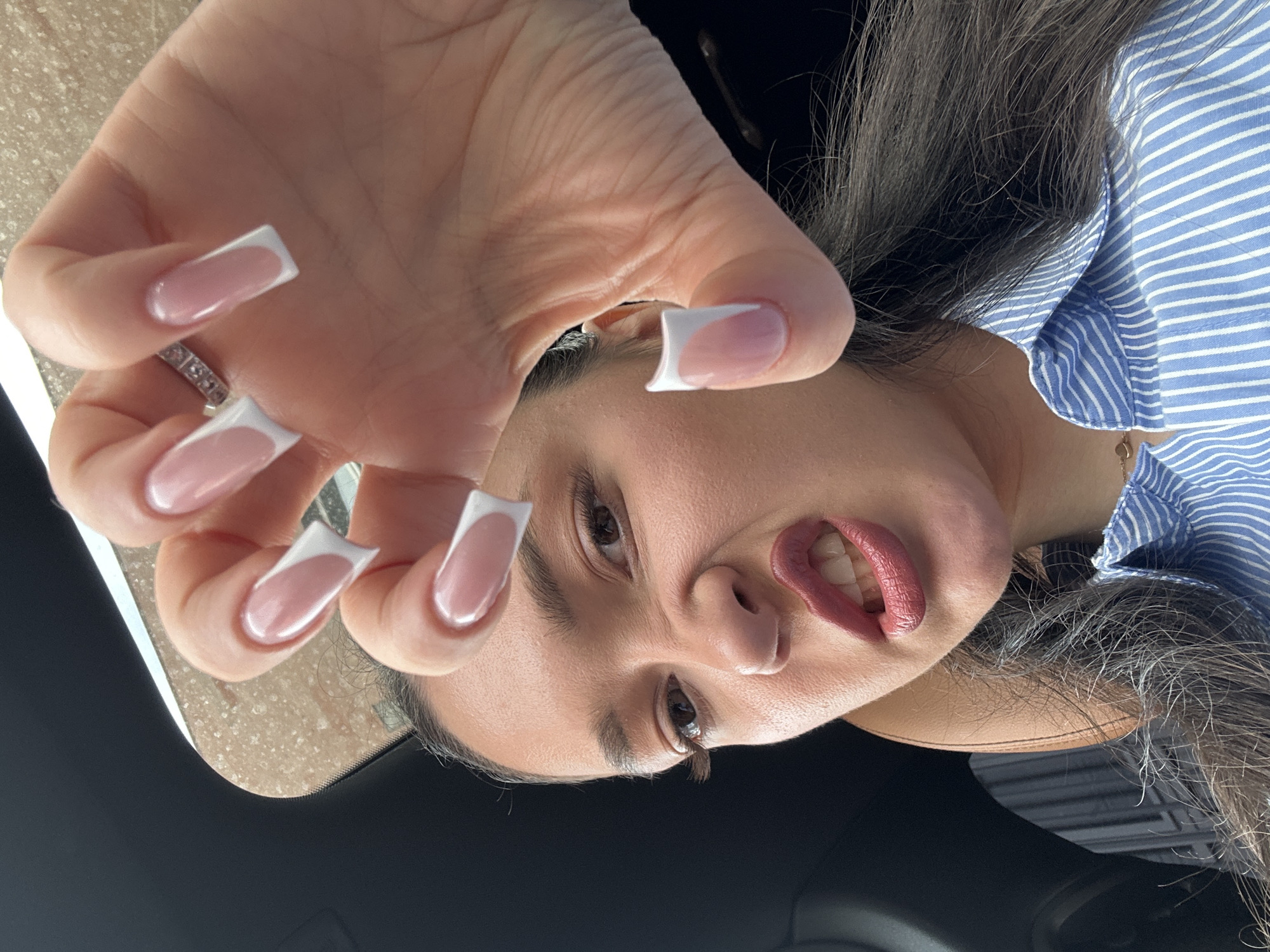 A person is facing the camera, with their hand in the foreground displaying French-tip manicured nails. The person has dark hair and light brown eyes. They are wearing a blue and white striped shirt and are likely seated in a car. The background is blurry, suggesting an urban environment. It's impossible to pinpoint the specific location within Yerevan, Armenia, without additional context or visible landmarks.