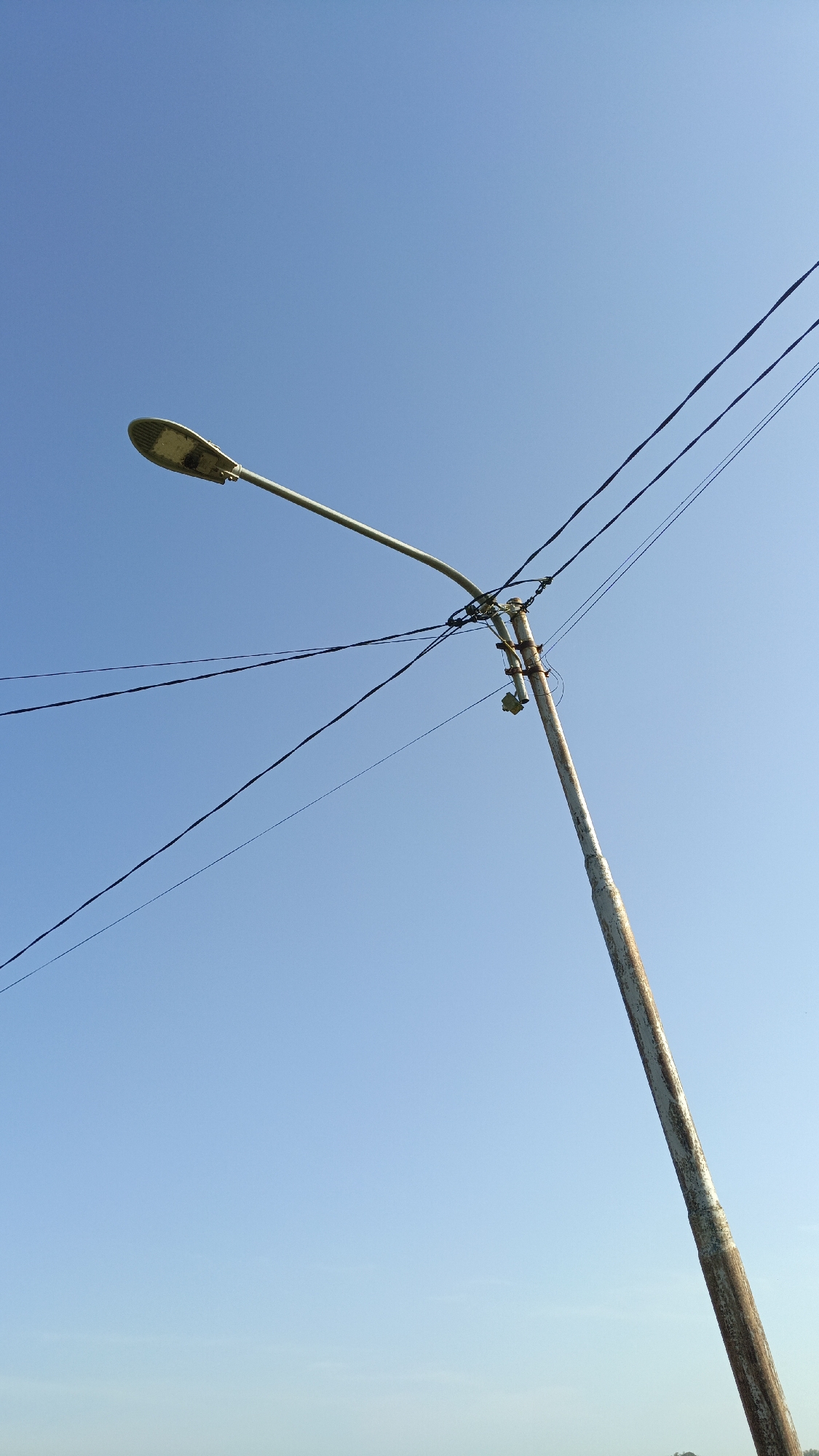 In Batarang, Indonesia, a tall, weathered street lamp post stands against a clear blue sky. The post, which appears to be made of metal and shows signs of rust and wear, supports a street lamp at its apex. Multiple black electrical wires are strung from the post, crossing the sky in various directions. The street lamp itself is elongated and grey, with its light-emitting surface facing downwards. The scene is set outdoors, with no other significant structures or individuals visible in the frame. The lighting suggests it is daytime, with the sun likely positioned overhead, casting no discernible shadows from the main subject.