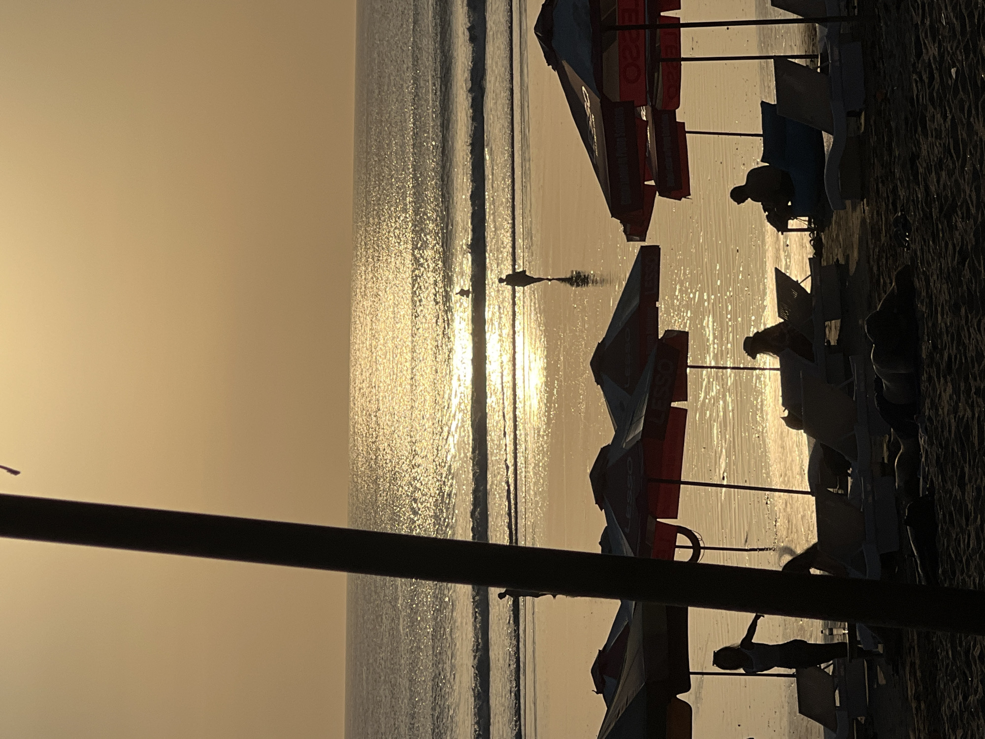 A silhouette of a person stands on a wet sandy beach facing the ocean at sunset. The sun's reflection glints off the calm ocean water. Several beach umbrellas are set up on the sand in the foreground, with lounge chairs visible underneath them. Silhouettes of people can be seen sitting in some of the chairs. The image captures a serene beach scene in Kuta, Indonesia.