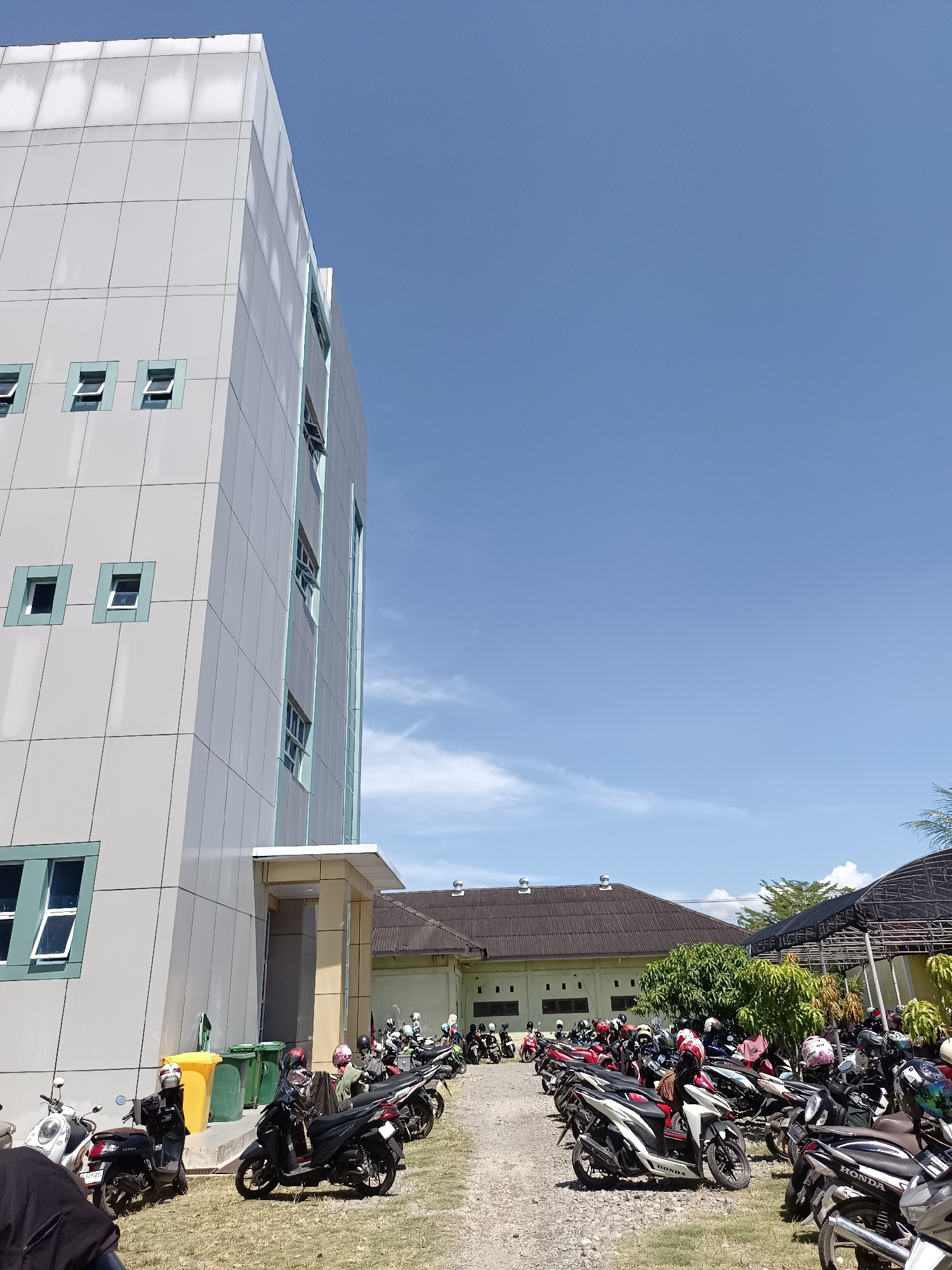 This is a low-angle, wide shot of a modern, light-colored building on the left, with a bright blue sky and scattered clouds above. The building has a grid-like facade with several small, square windows, some of which are open. To the right of the building is a parking lot filled with many parked motorcycles and scooters. The motorcycles are of various colors, with several individuals wearing helmets parked or sitting on them. A few people are visible in the parking lot, though their faces are not clearly discernible. In the background, there is a lower building with a dark roof and some trees. The scene appears to be on a sunny day. The image was taken in the city of Mesjid Reuntoh, Indonesia.