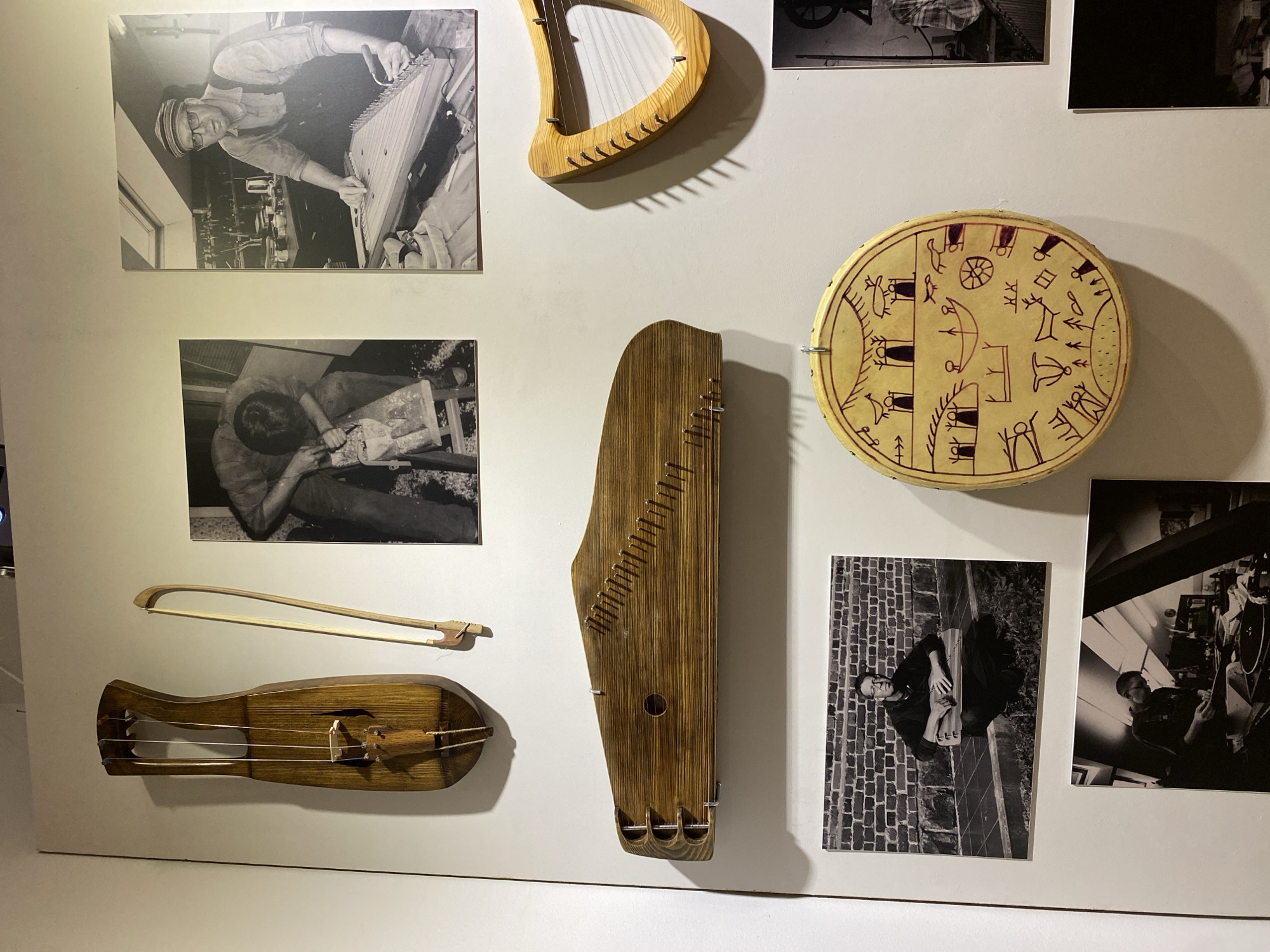 The image displays a wall in a museum exhibiting various musical instruments and photographs. The museum is located in Petrozavodsk, Russia.
The wall is white and serves as a backdrop for the displayed items.
At the top left, a wooden stringed instrument resembling a kantele or lyre with a long, curved body and three strings is mounted vertically. To its right is a wooden bow.
To the right of these instruments are two black and white photographs. The first shows a person carving wood with a tool, with shavings scattered around. The second photograph depicts a man wearing glasses and a cap, with overalls, working on a large, flat stringed instrument with numerous strings.
Below the first set of instruments, a long, wooden stringed instrument with many strings is mounted horizontally across the wall. It has a curved shape with tuning pegs.
To the right of this horizontal instrument is a smaller, harp-like wooden instrument with strings.
Further down, there are more photographs. A black and white photograph shows a man sitting outside in front of a brick wall, holding a stringed instrument. Below that, another black and white photograph shows a person working on a musical instrument in an indoor workshop with slanted ceilings.
In the center of the lower portion of the wall, a circular drum with intricate red drawings on its surface is hung. The drawings depict various figures and symbols, possibly representing a shamanic or folk theme.
The overall scene is that of a museum display, showcasing traditional musical instruments and imagery related to their creation and use.