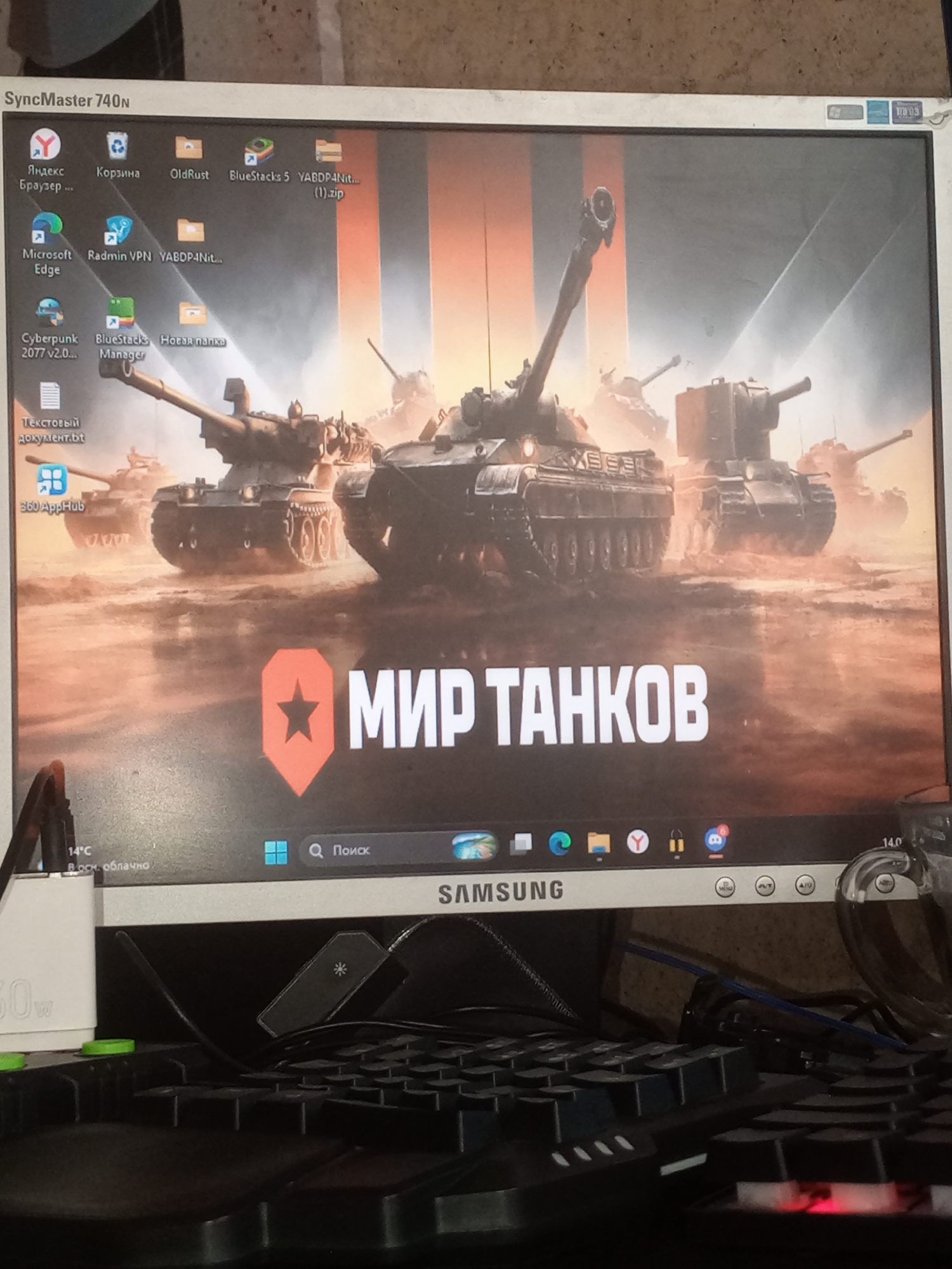 The image shows a computer monitor displaying a desktop wallpaper with the title "МИР ТАНКОВ" (World of Tanks) in large white letters. The wallpaper features an image of several tanks on a battlefield, possibly in a desert or dusty environment. On the left side of the screen, there are several desktop icons, including Yandex Browser, Recycle Bin, OldRust, Bluestacks 5, and various folders and files.
The monitor is a Samsung SyncMaster 740N. In the lower part of the image, a keyboard is visible, along with some cables and a white power adapter. The overall environment appears to be a room, likely a home or office setting, with a textured wall in the background. There are no people visible in the image.
The image is taken from an angle, suggesting it was captured by a person using a camera or smartphone to photograph the computer screen. There are no elements in the image that can be used to determine its specific location in Krasnokamensk, Russia.