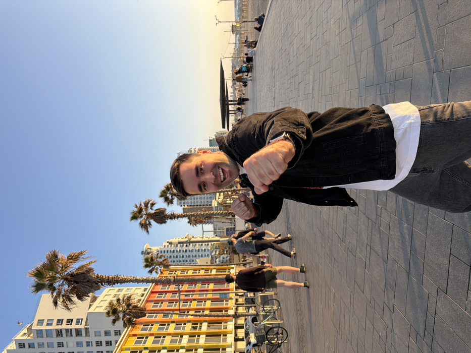 The image captures an outdoor scene on the Tel-Aviv Promenade (Tayelet) in Tel-Aviv, Israel, during late afternoon or early evening. The main subject is a light-skinned male in his late 20s to early 30s, with short dark hair, centered slightly to the right of the frame. He is wearing a black denim jacket over a white t-shirt and dark grey jeans. He faces the camera directly, displaying a wide smile with an open mouth, and has both fists raised in a simulated boxing or fighting pose. A silver-colored watch is visible on his left wrist.
The ground consists of light grey rectangular paving stones forming the promenade. To the left, a row of multi-story buildings, including one with distinctive colored facade panels (orange, yellow, green, red, blue), stands behind several tall palm trees. Further back, more high-rise buildings are visible. Multiple individuals are walking on the promenade in the background, and two bicycles are parked near the left edge.
To the right of the promenade, a sandy beach extends, populated by several people sitting or walking. Beach structures, possibly shaded seating or lifeguard posts, are visible on the sand. The sky is clear, transitioning from light blue at the zenith to warmer orange and yellow tones near the horizon, consistent with a sunset.