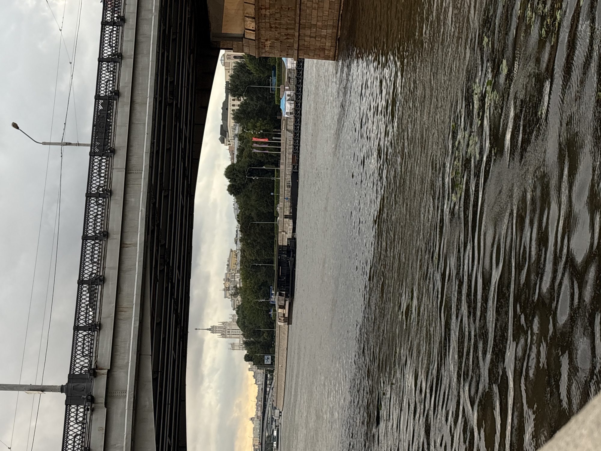 This image captures a view from the Moskva River in Moscow, Russia, looking towards the Stalinist skyscraper, Kudrinskaya Ploshchad, which dominates the skyline. A large, multi-level bridge spans the frame from the top, obscuring part of the view. Beneath the bridge, a black boat or barge is visible on the river, moving away from the viewer. The riverbank features a stone embankment and a line of trees. In the background, beyond the trees, several buildings are visible, including the prominent Kudrinskaya Ploshchad. The sky is overcast, with clouds suggesting a potentially grey or rainy day. The water in the foreground is rippled, showing small patches of green aquatic plants. The overall scene depicts a city waterfront on a cloudy day, with significant architectural structures and watercraft present.