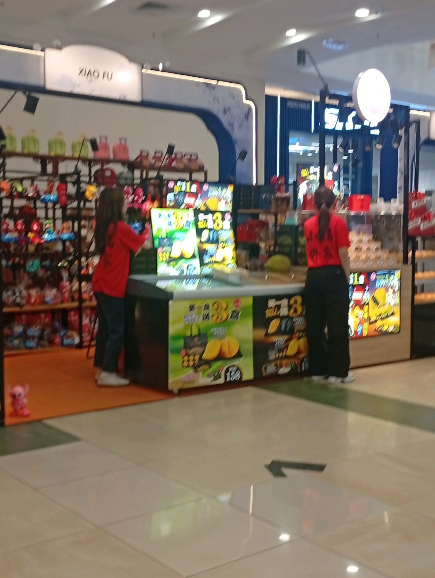 The image shows an indoor marketplace or shop. A sign above a display reads "XIAO FU". Two women are visible. One woman, wearing a red t-shirt and dark pants, is standing in front of a display of food items. She is interacting with the display. The other woman, also in a red t-shirt and dark pants, is standing further back and facing away from the camera. The shop appears to be selling various items, with shelves full of products in the background. The floor is tiled, and a directional arrow is visible on the floor in the foreground. The lighting suggests an indoor commercial setting. The image does not provide enough information to determine the specific location within Kuala Lumpur, Malaysia.