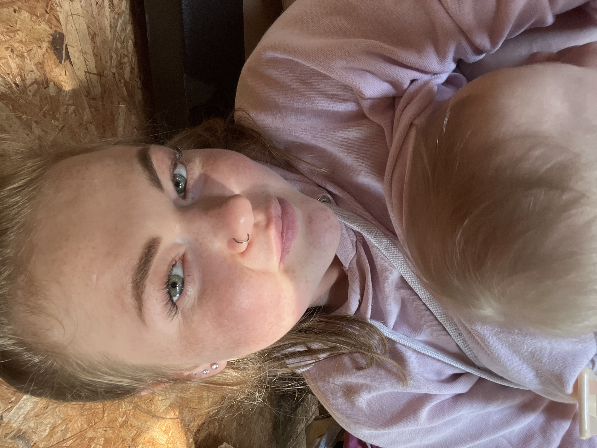 The media file is a close-up, portrait-style photograph of a young woman lying down. She is wearing a light pink hooded sweatshirt. Her head is tilted, and she is looking towards the camera with a soft expression. She has light brown hair, fair skin with freckles, and green eyes. She is wearing a small silver hoop in her nose and two small stud earrings on her right ear.
The background appears to be made of unfinished wood, possibly a floor or wall, with some texture and variations in color. The lighting suggests it might be daytime, with natural light casting shadows on her face. The overall mood is casual and intimate. No text is visible in the image. The location context is Chaikovsky, Russia.