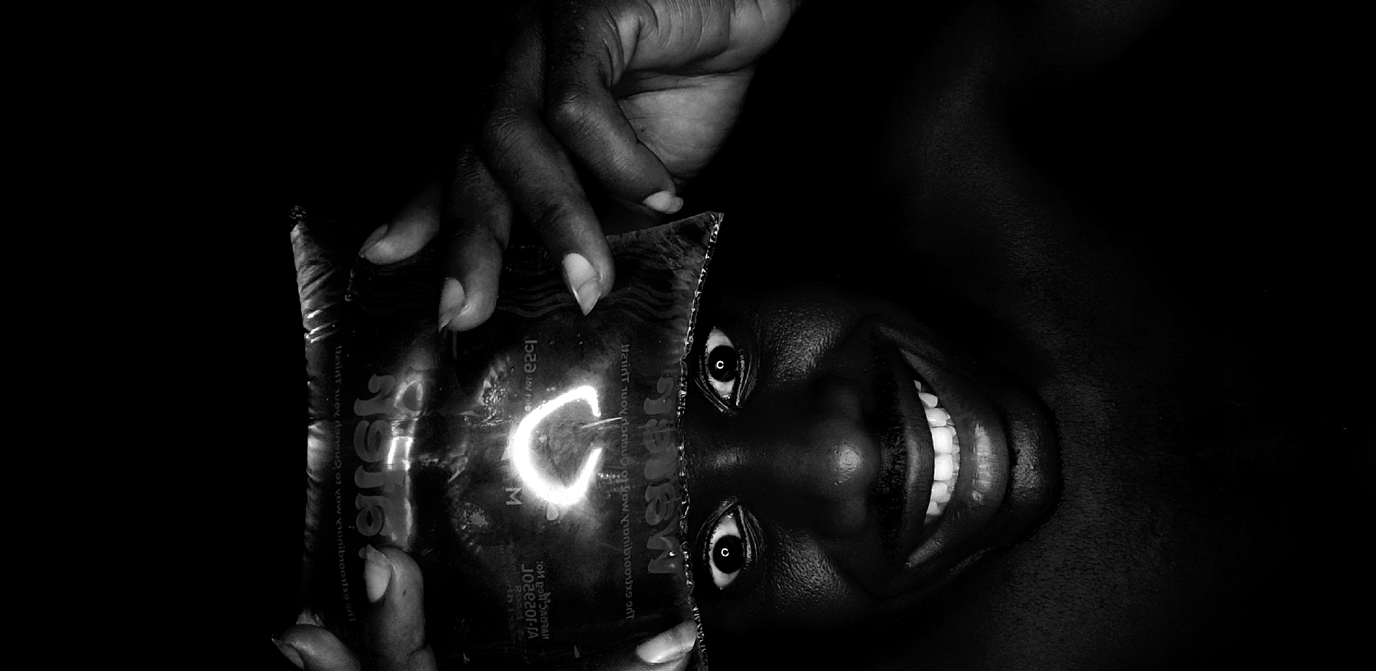 A black and white, high-contrast image shows an upside-down view of a dark-skinned person. The person is smiling widely, exposing their teeth. A translucent, shiny package bearing the word "Matrix" in large letters is held close to the face, obscuring part of the forehead. The person's eyes are visible through the package. A hand is holding the package. The background is dark. No specific location is identifiable in the image.