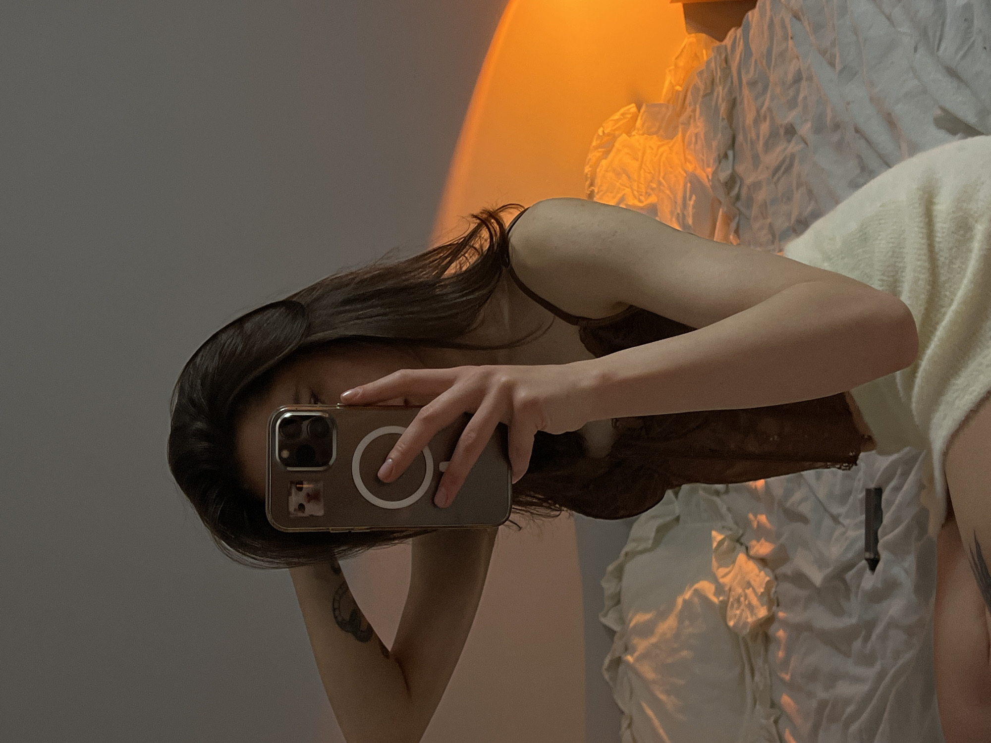 The image shows a young woman in her bedroom, taking a mirror selfie. She is wearing a brown lace camisole and a white fuzzy skirt. Her right arm is raised, touching her head, while her left hand holds up a phone to take the photo. The phone has a light brown case with a circular magnetic charger visible on the back. A small image of a cat is visible on the phone's case. A tattoo is visible on her left forearm.
The bedroom is dimly lit, with warm orange light casting a glow on the wall and bed. The bed is covered with white sheets, which appear slightly rumpled. A crumpled piece of white fabric is on the bed near the center, and a dark rectangular object, possibly a pen or a remote, is also on the bed.
The image does not contain any information that can be used to determine its location in the city of Ternopil, Ukraine.