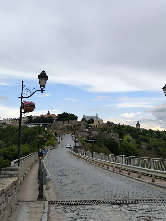 The image depicts a longitudinal view of the Novoplanivskyi Bridge in Kamianets-Podilskyi, Ukraine, extending northward towards the city's Old Town. The scene is illuminated by diffused daylight under a sky displaying a mixture of light grey cloud cover and patches of blue.
In the immediate foreground, a cobblestone road surface features two parallel rectangular metal storm drain grates. To the far left, a low stone wall forms the edge of a pedestrian walkway. Positioned on this walkway is a prominent black, ornate lamppost, approximately 3 meters in height, from which a terracotta pot containing pink flowering plants is suspended. Adjacent to this lamppost, a metal barrier with vertical slats delineates the pedestrian path from the main vehicular bridge roadway.
Two individuals, identified as females, are observed walking away from the viewer on this left pedestrian path. The bridge roadway, composed entirely of cobblestones, gently ascends and curves slightly to the left as it proceeds towards the elevated city. Metal guardrails with vertical slats line both sides of the bridge's vehicular lanes. Additional black, ornate lampposts with suspended flower pots are spaced along the entire visible length of the bridge on both sides.
Approximately eight to ten additional individuals are visible as pedestrians on both the left and right bridge walkways in the mid-ground, walking in various directions. A single white passenger vehicle is present on the cobblestone bridge roadway, proceeding northward towards the city. Several red passenger vehicles are parked on the right side of the road further along the bridge, approximately 50 meters from the foreground.
The background reveals the elevated historic Old Town of Kamianets-Podilskyi, characterized by extensive stone fortifications, including walls and remnants of turrets. Multiple historic buildings are visible, including a light-colored structure with a distinct roofline (likely a church or monastery) and a distant steeple with a dark, conical roof, both situated atop the elevated land. Dense green foliage, primarily trees and bushes, covers the natural slopes and ravines surrounding the bridge and the base of the historic city structures.