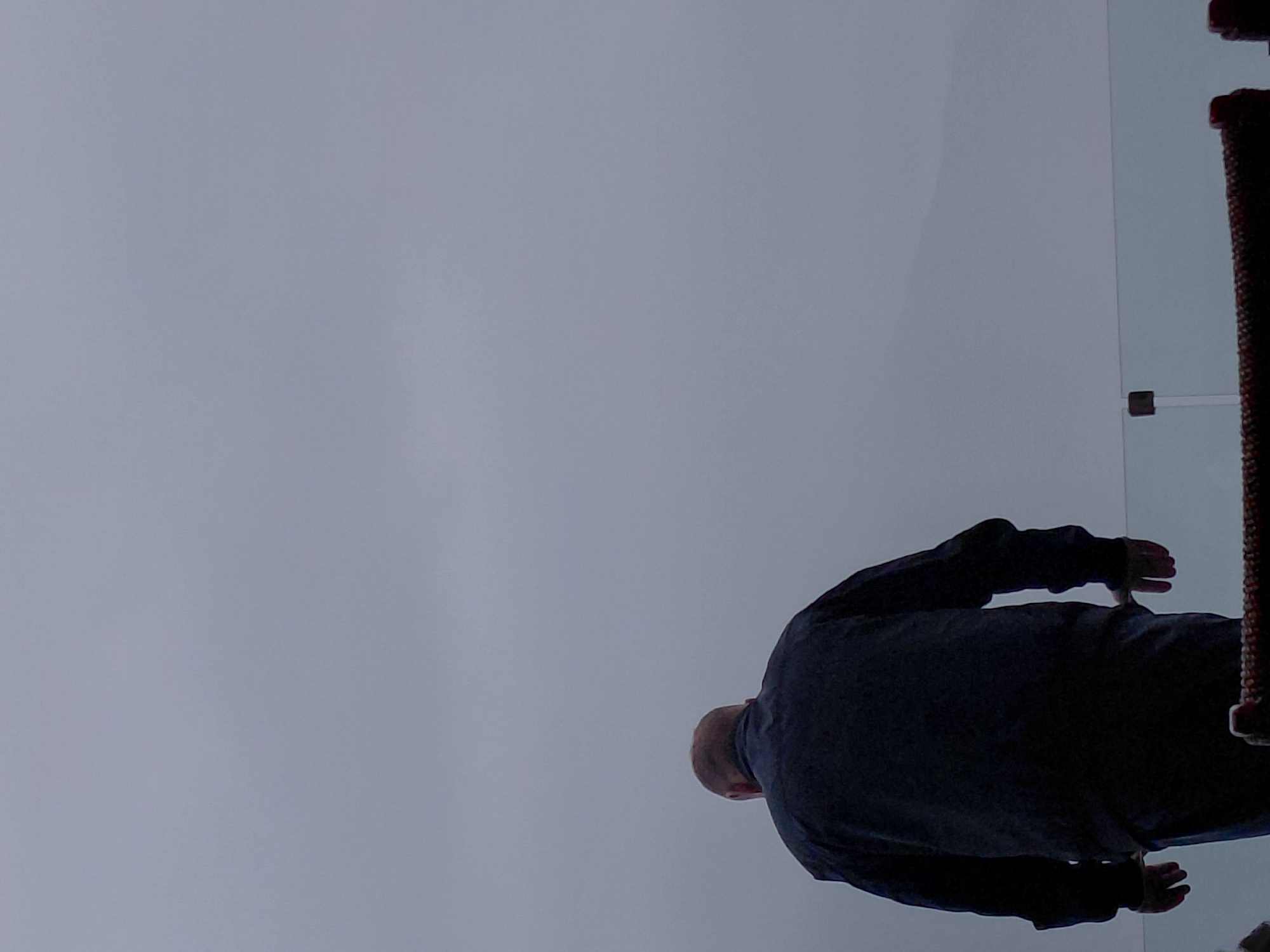 A person wearing a dark jacket and blue jeans stands with their back to the camera, looking down. The person is in the foreground, with a blurred background of an overcast sky and distant hills. A glass railing is visible in front of the person, and a portion of a railing with a patterned texture is also in view at the bottom of the image.
