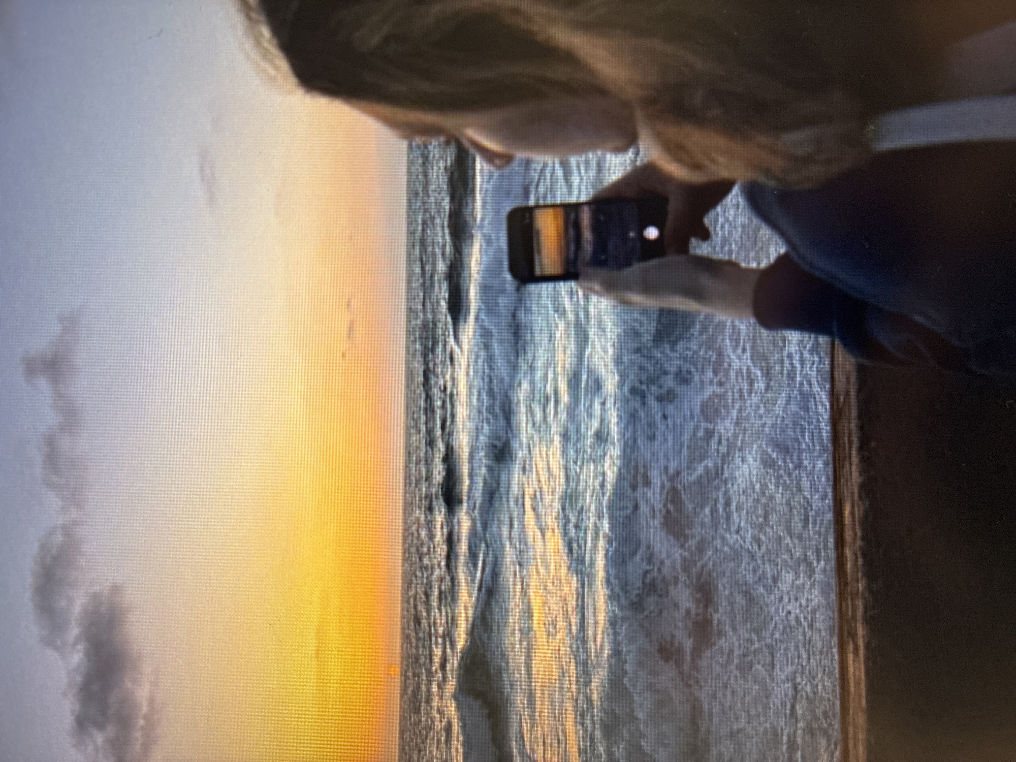A person is holding a smartphone to photograph a sunset over the ocean. The sky transitions from gray on the left to orange towards the center of the frame. Waves are visible in the ocean. This is likely a scene in Bat Yam, Israel due to the coastal location.