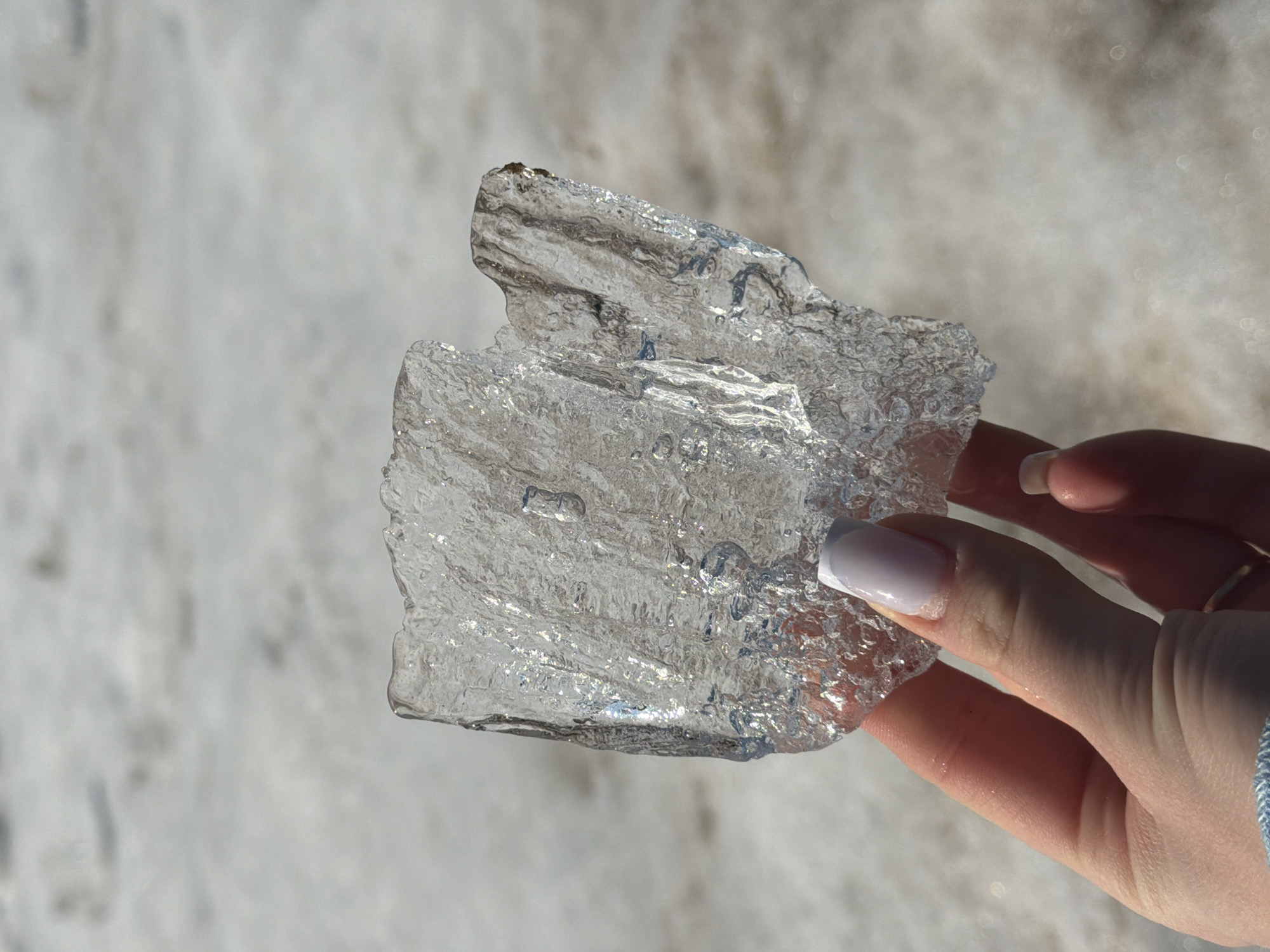 A person is holding a piece of clear ice in their right hand. The ice is roughly square with uneven edges and internal refractions. The person's fingers are visible, with a pale manicure on the nail of the index finger. A gold ring is on the ring finger, and a blue and white cord is wrapped around the wrist. The background is out of focus and appears to be snow-covered ground. The location is not discernible from the image. The city of Chaikovsky, Russia, cannot be determined from the visual information.