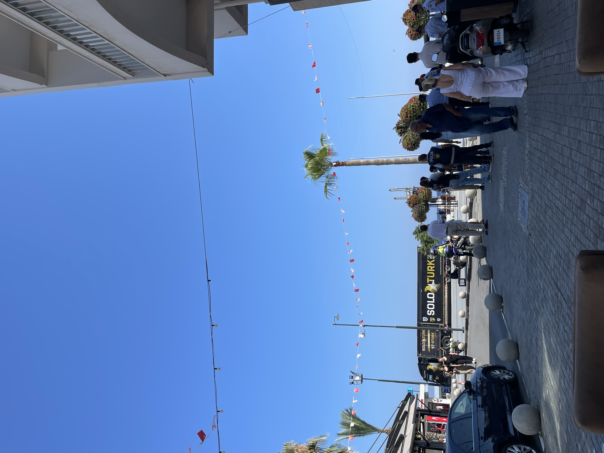 A street scene in Kyrenia, Northern Cyprus, features a clear blue sky with strings of red and white flags and lights decorating the area. The street is paved with interlocking bricks, and large, spherical concrete bollards line the walkway. On the left, a dark-colored car is parked, and a building with shops is visible. A black truck with "SOLO TURK" branding is parked further down the street, alongside a motorcycle. A group of people, some in what appear to be police uniforms, are gathered on the sidewalk, some conversing, others standing or walking. A tall palm tree stands prominently in the middle ground. In the background, the sea and port infrastructure are visible. The architecture suggests a coastal Mediterranean setting.